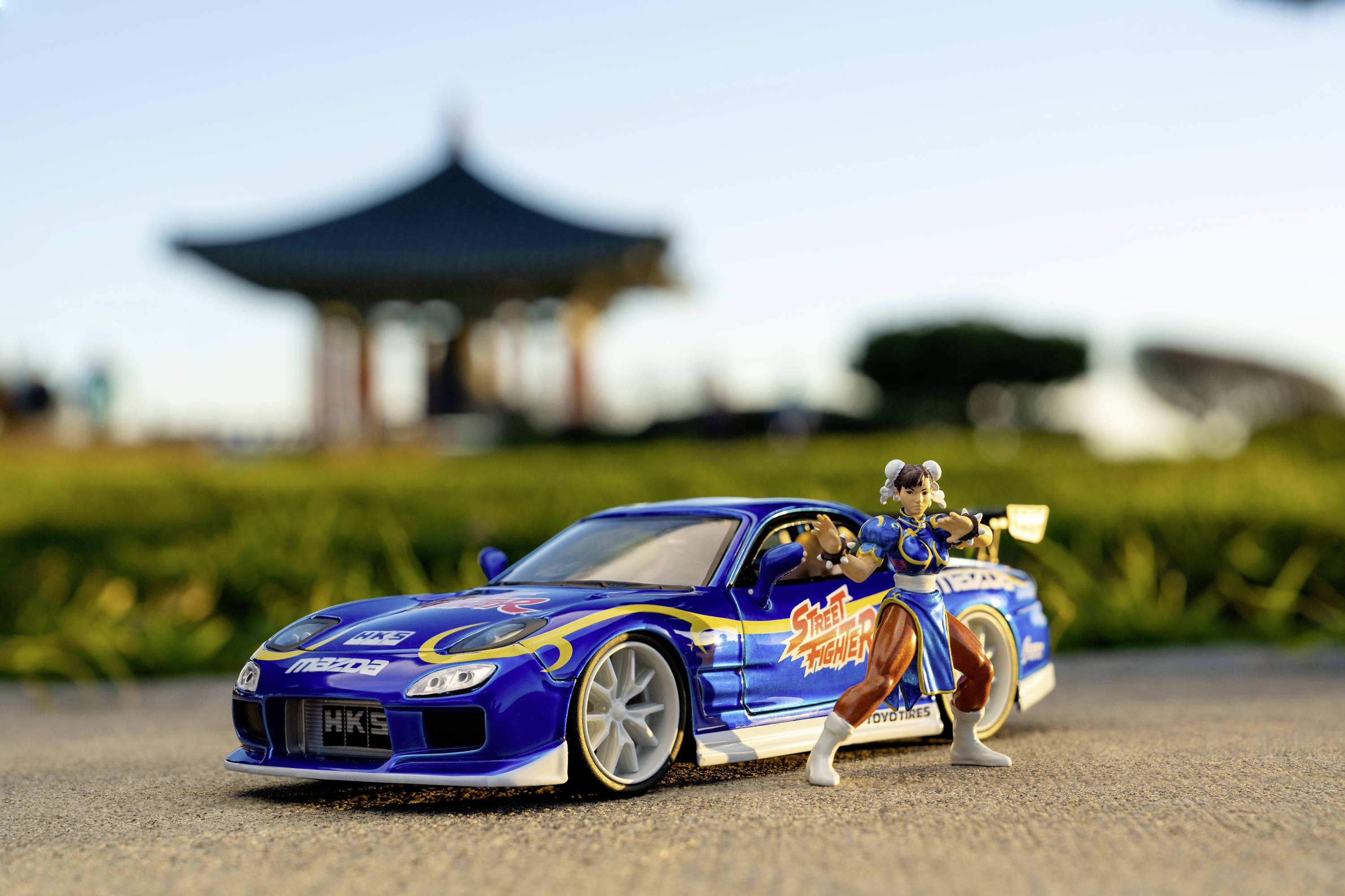 Modrý model závodního auta s nálepkami 'Street Racer', vedle něj malá bojová figurka. V pozadí pavilon a stromy.
