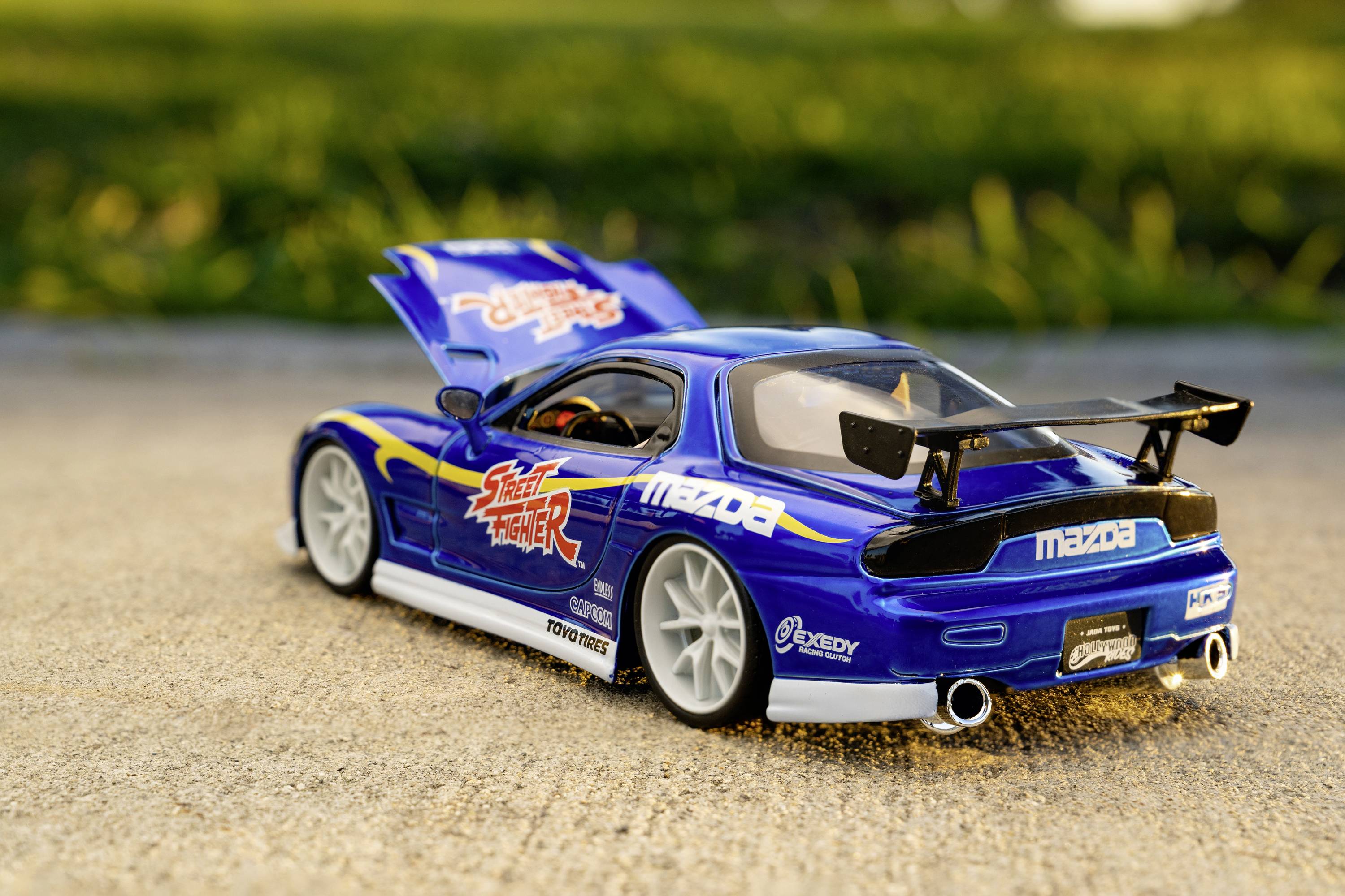 Modrý modelový sportovní vůz s otevřeným zavazadlovým prostorem stojí na betonu před zeleným pozadím. Auto má spoiler a nálepky.