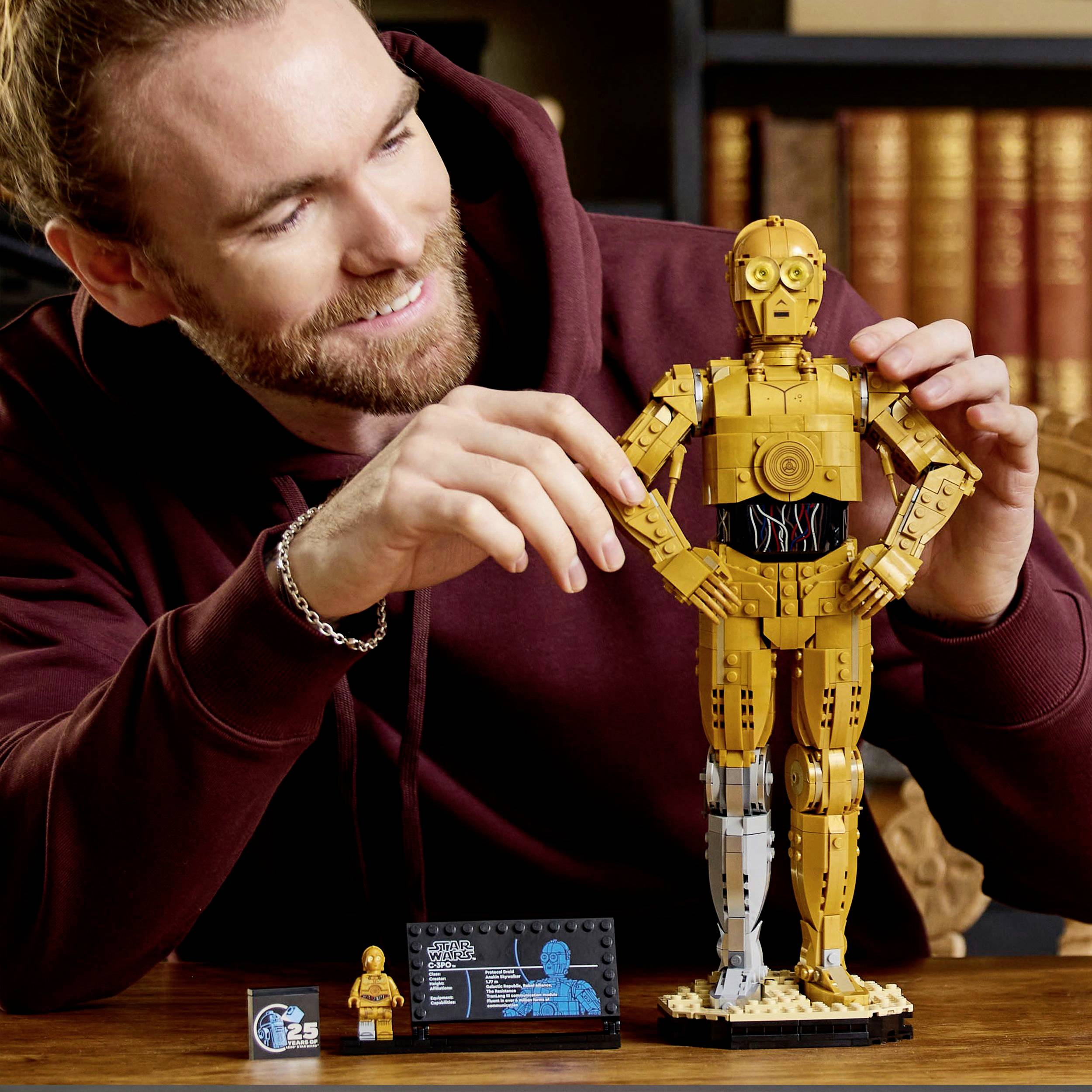 Osoba v mikině s kapucí se usmívá a dívá se na detailní model humanoidního robota umístěného na stole.