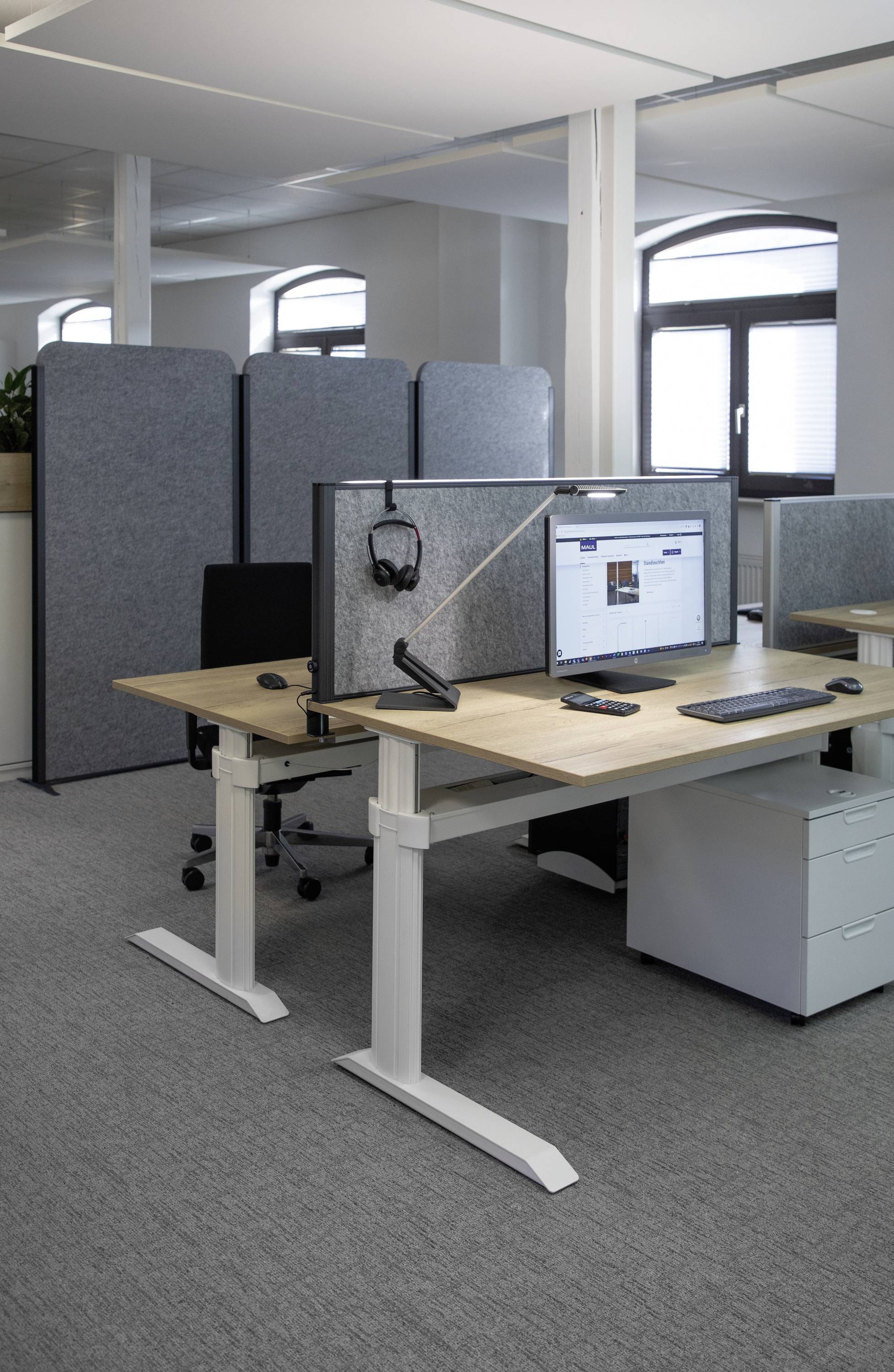 Moderní kancelář s výškově nastavitelnými psacími stoly, monitory a laptopy. Zástěny oddělují jednotlivá pracovní místa.
