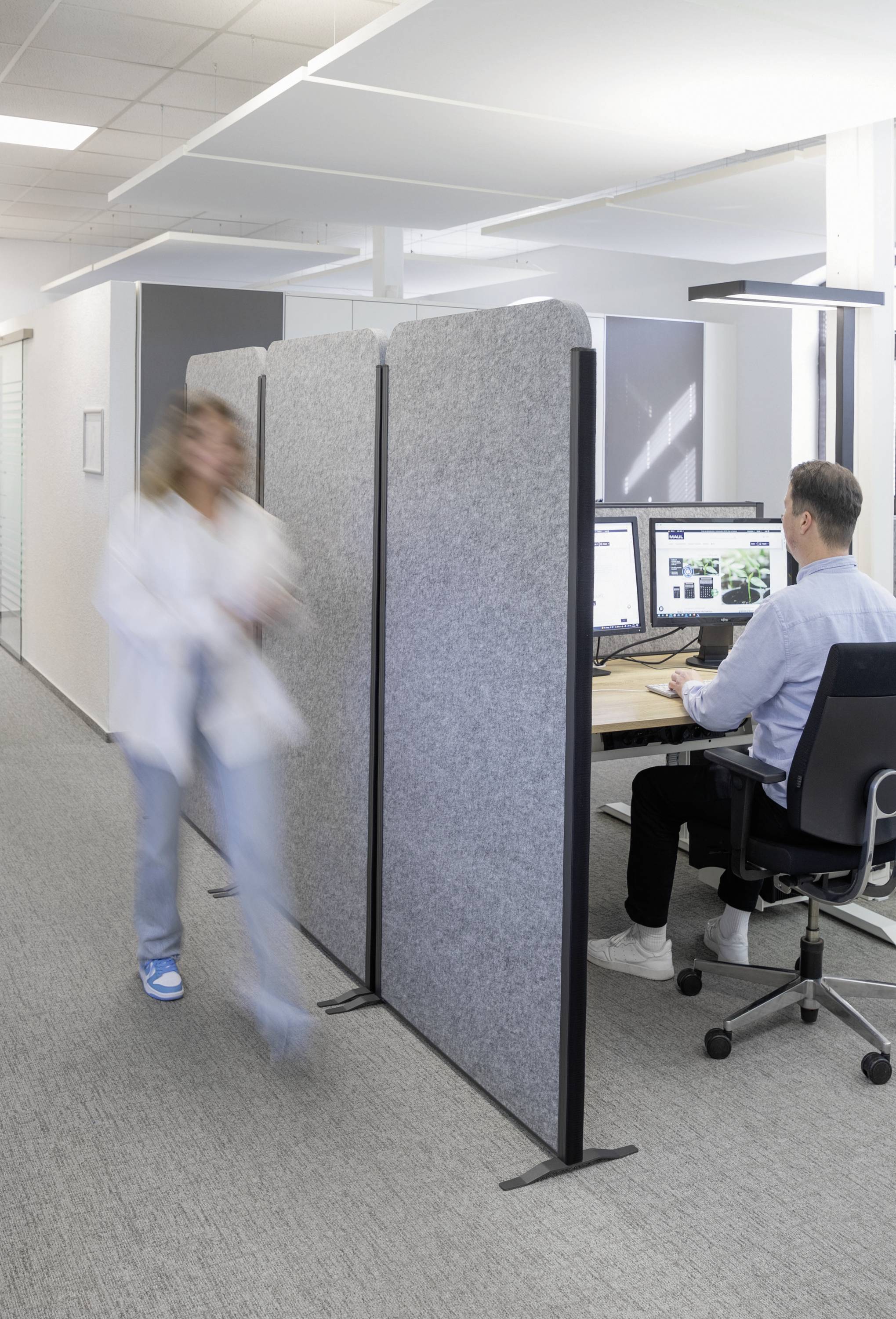 Muž sedí u psacího stolu a pracuje na počítači. Žena rychle prochází kolem. Kancelář s moderním designem a přepážkami.