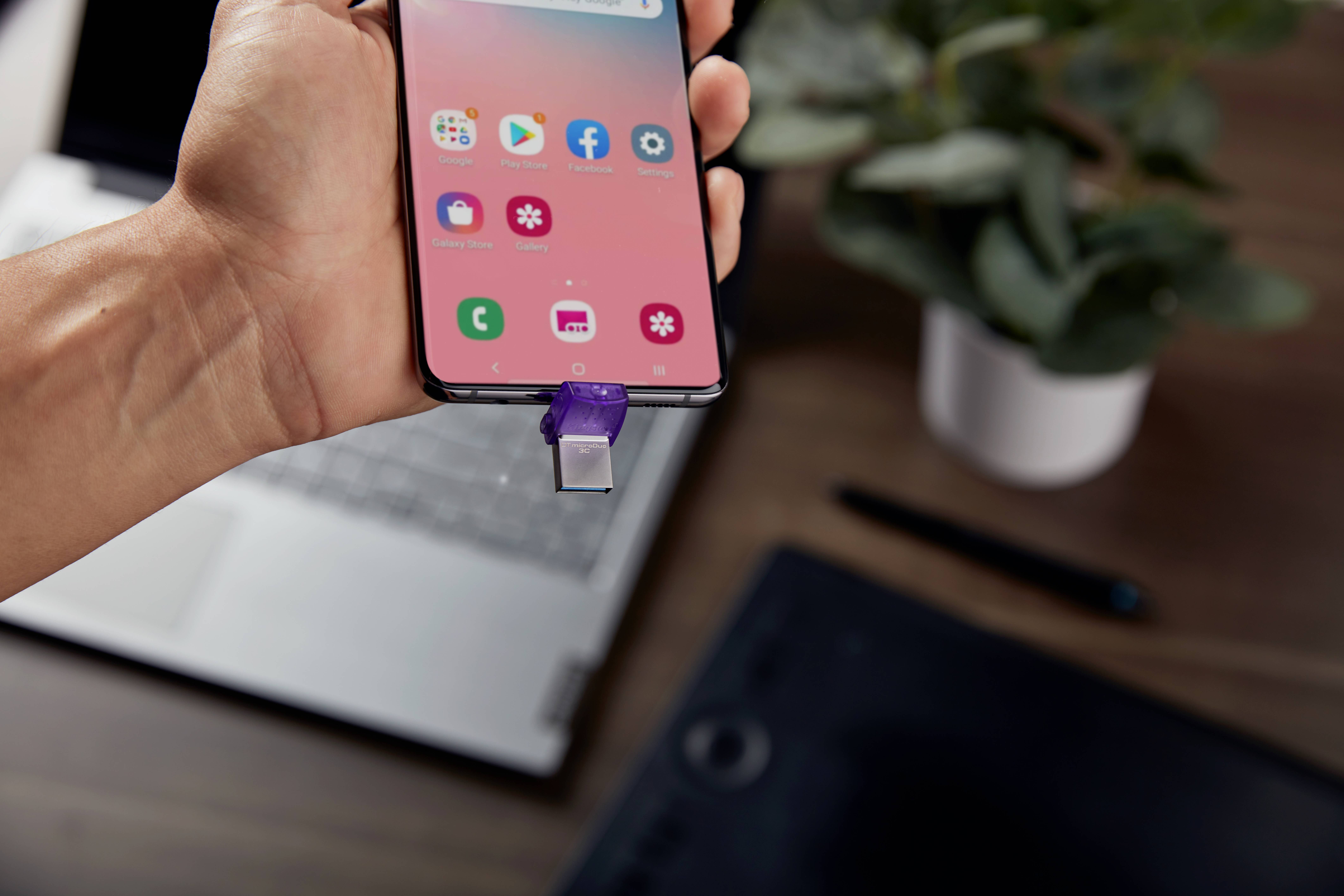 Jedna ruka drží smartphone, ke kterému je připojena USB flash disk. V pozadí je notebook, pero a rostlina.