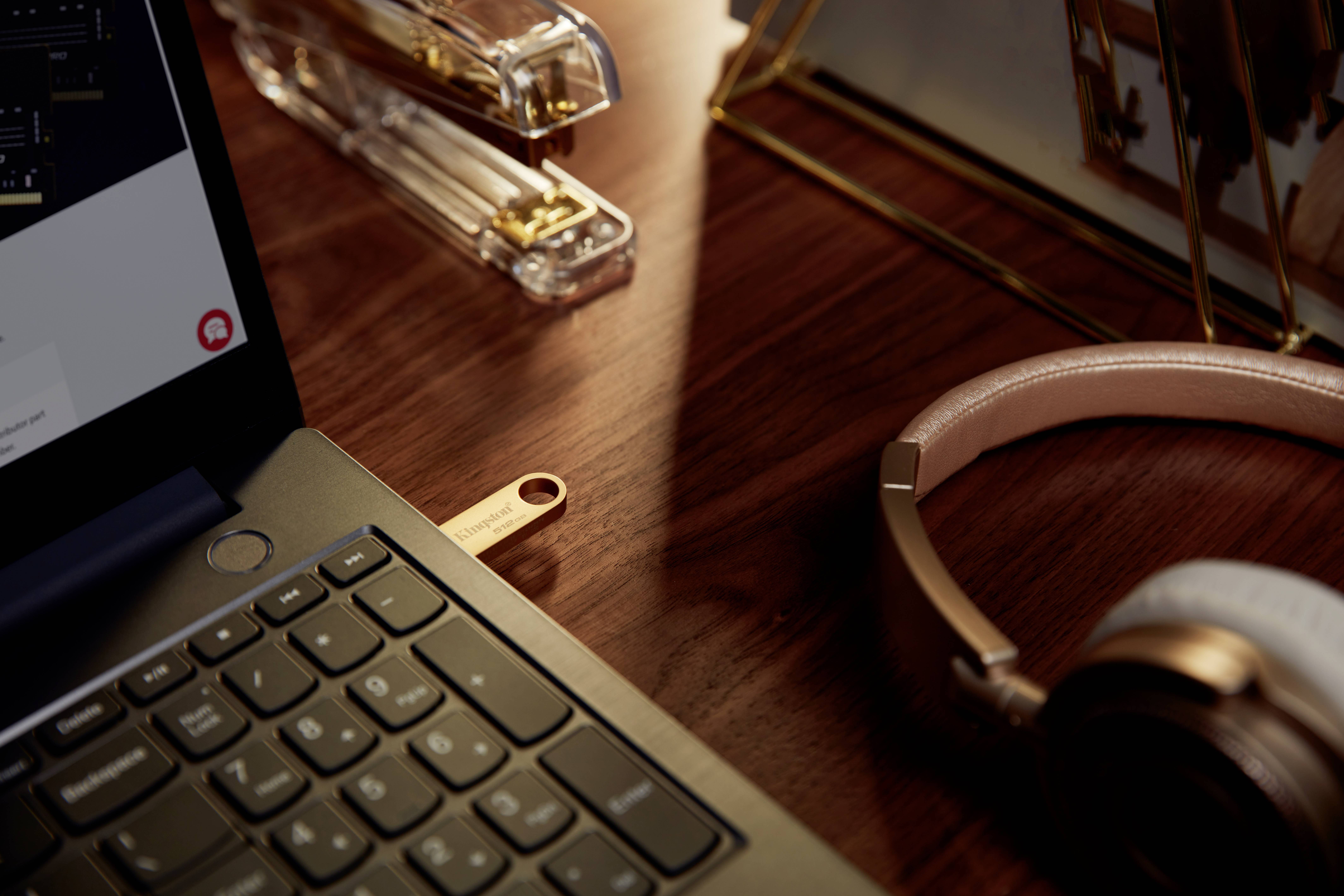 Laptop na dřevěném stole s USB flash diskem, vedle sluchátka a zásobník na kancelářské sponky. Atmosféra: útulné pracovní prostředí.