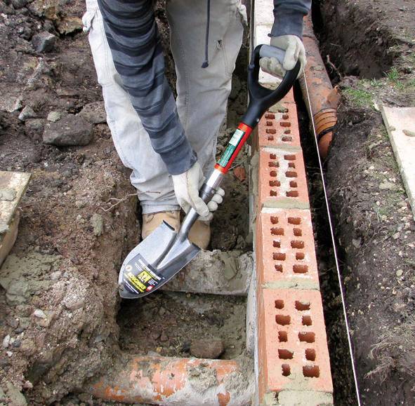 Osoba klade cihlové kameny v připravené řadě v zemi. Je viditelné vedení hadice. Činnost se vztahuje ke stavebním pracím.