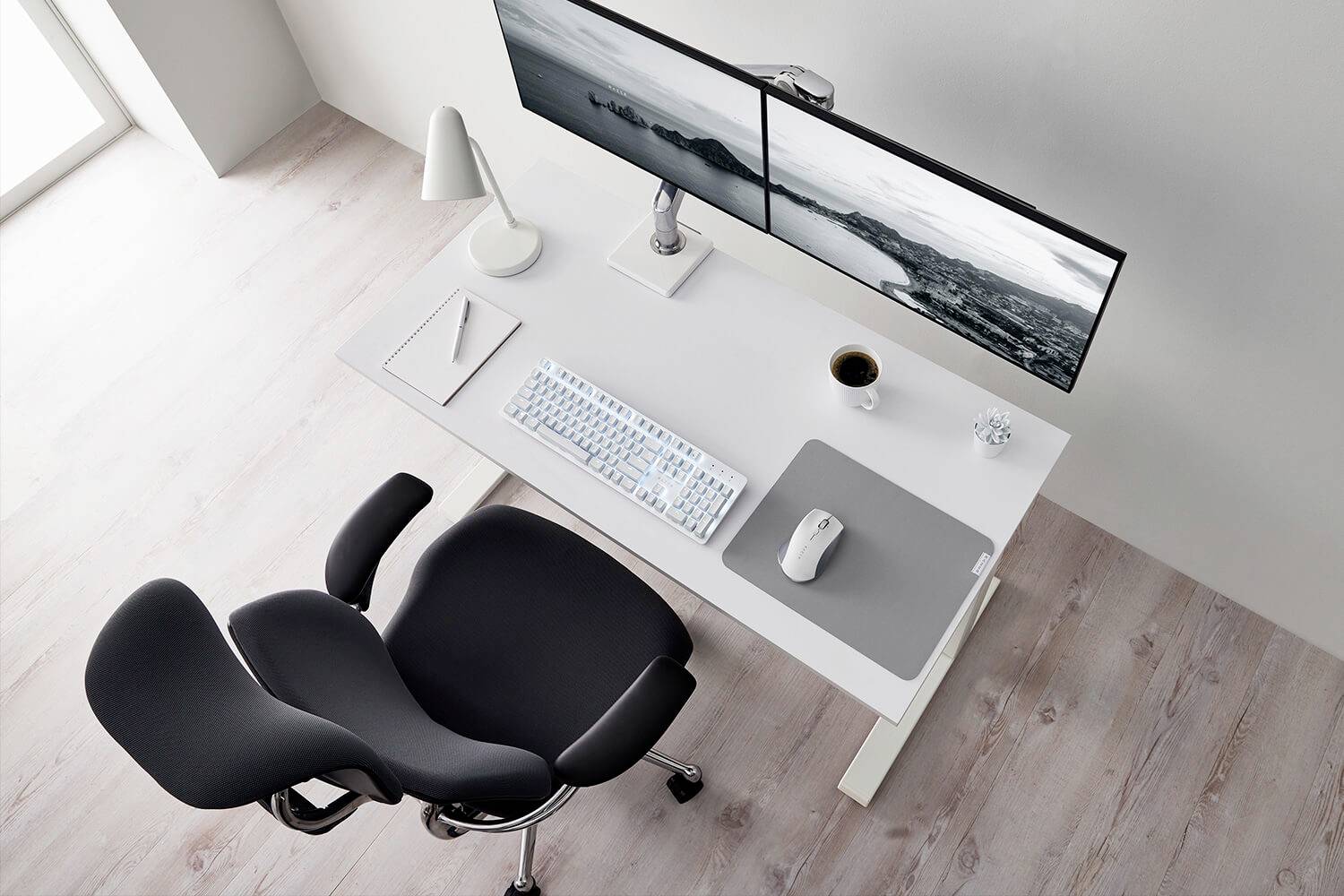 Moderní, uklizený psací stůl se dvěma monitory, klávesnicí, myší, poznámkovým blokem a šálkem kávy. Před ním stojí ergonomická židle.
