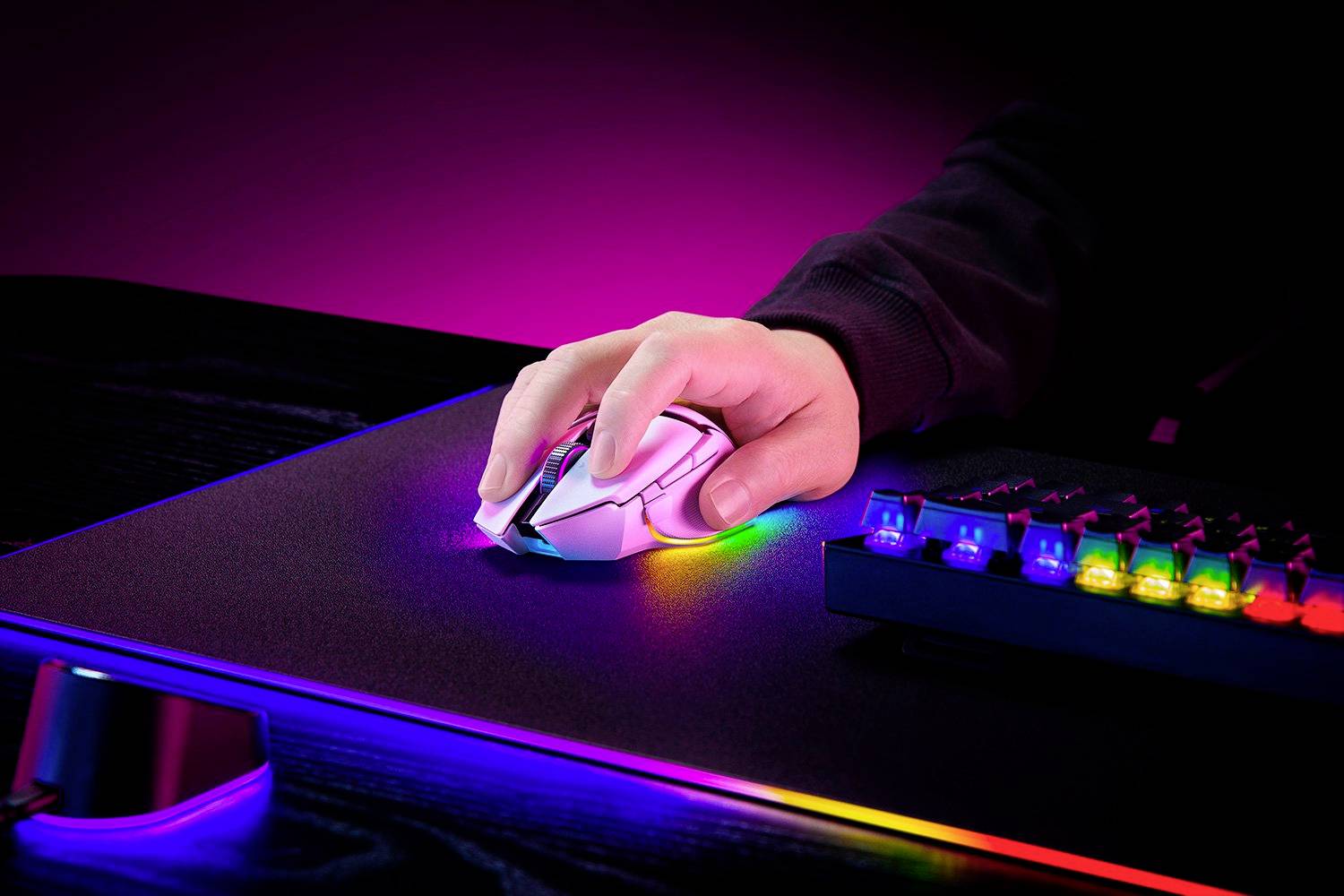 Ruka pohybuje svítící herní myší na podložce pod RGB osvětlením vedle klávesnice s barevnými klávesami.