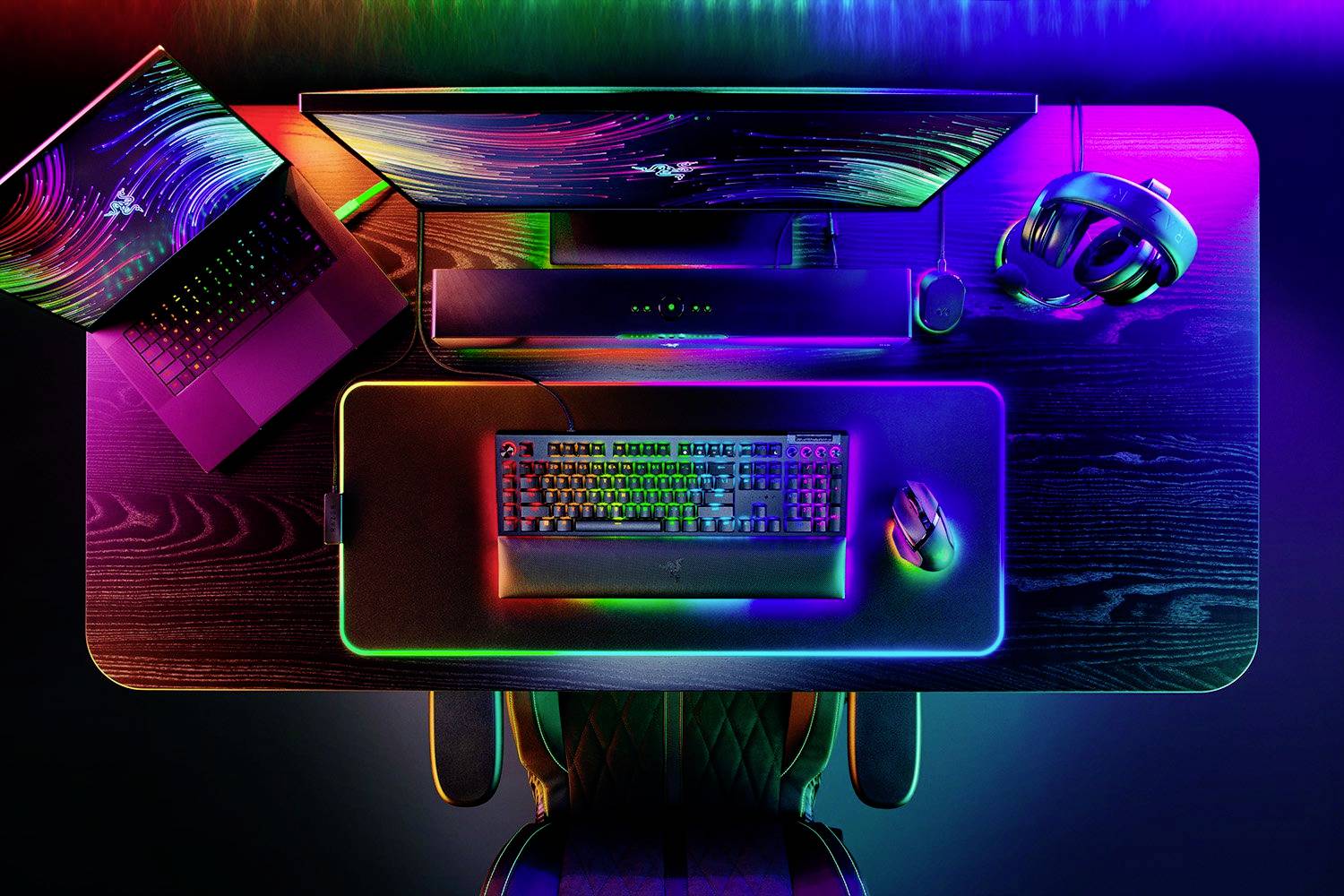 Psací stůl s barevně osvětleným herním příslušenstvím: klávesnice, myš, sluchátka, monitor a notebook.