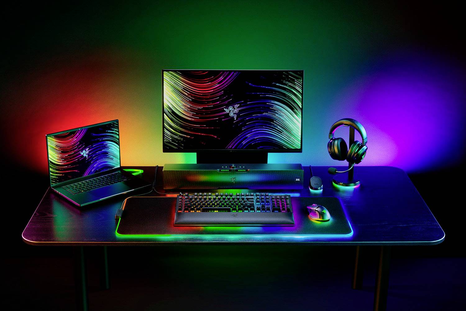 Psací stůl s laptopem, monitorem, klávesnicí, myší a sluchátky, všechny s barevným RGB osvětlením v tmavé místnosti.