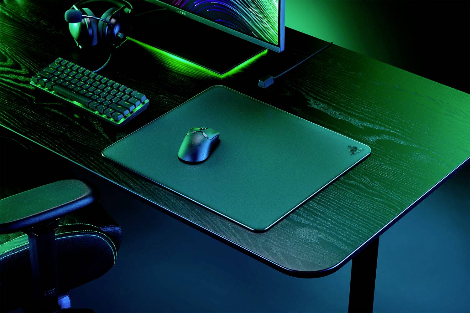 Černý počítačový stůl s klávesnicí, myší, podložkou pod myš a monitorem. Osvětlení v zelené barvě, herní sestava.