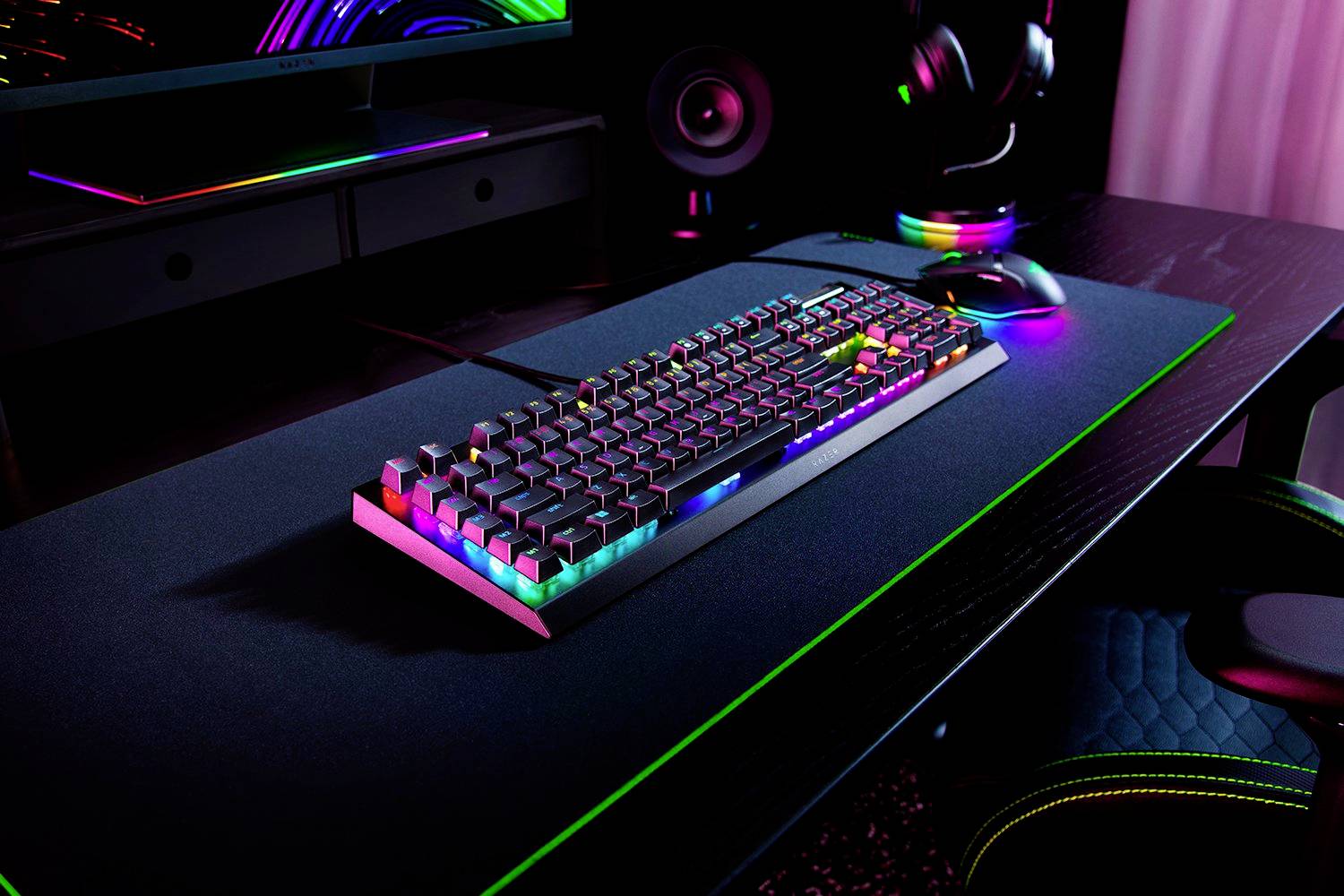RGB klávesnice na černém psacím stole, osvětlená v různých barvách, s počítačovou obrazovkou v pozadí.
