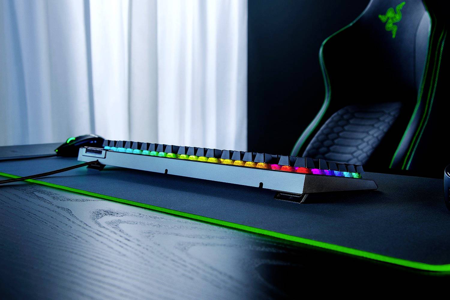 Herní klávesnice s barevným RGB podsvícením na psacím stole vedle herní myši a herního křesla.