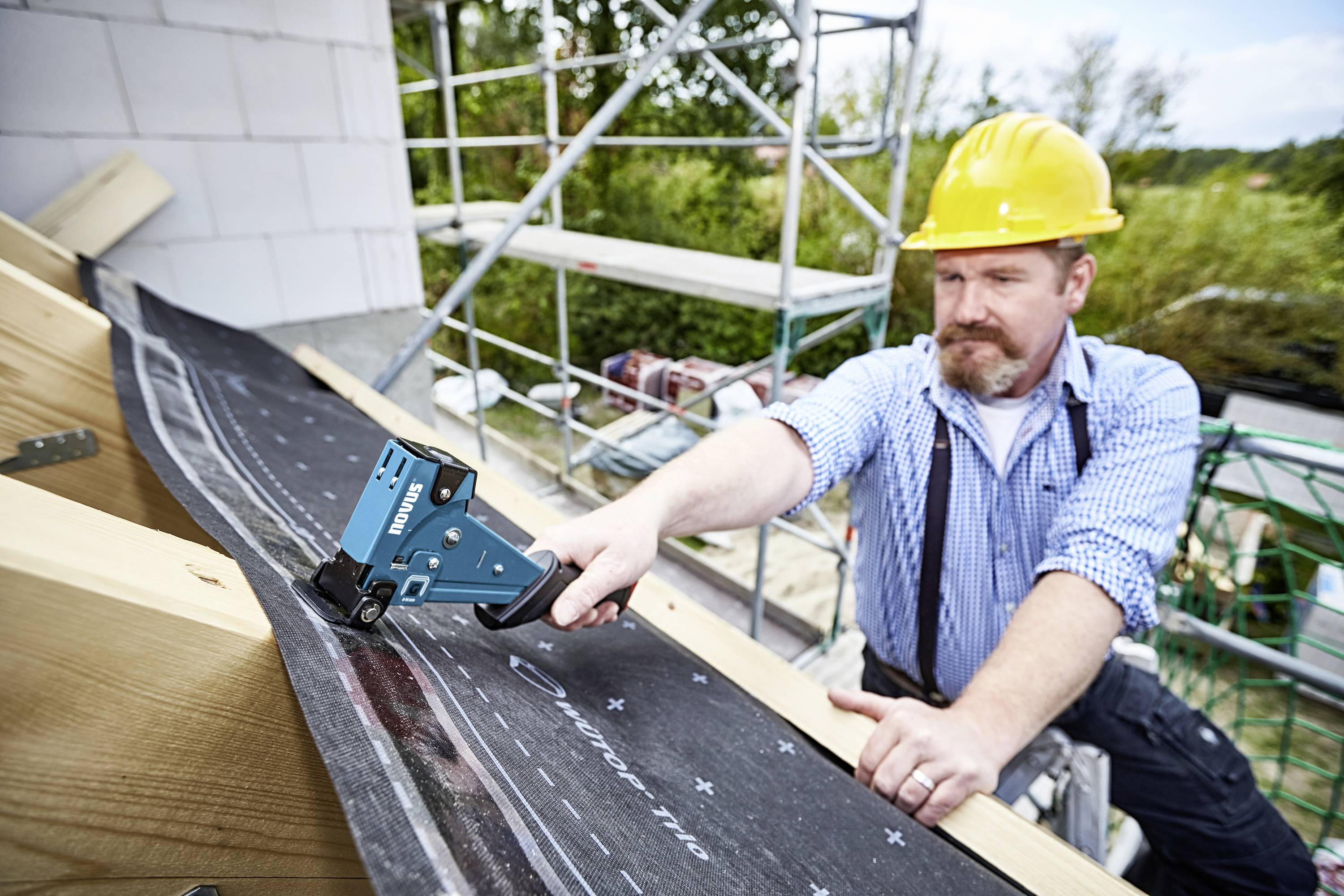 Dělník v žluté přilbě upevňuje střešní materiál nástrojem, zatímco stojí na lešení. V pozadí jsou vidět stromy.