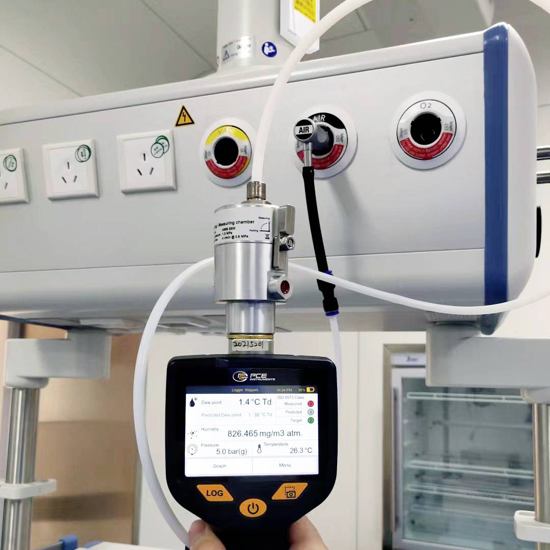 Ruka drží digitální měřič kvality vzduchu, který ukazuje 526,465 mg/m³ uprostřed místnosti s podrobným lékařským vybavením v pozadí.