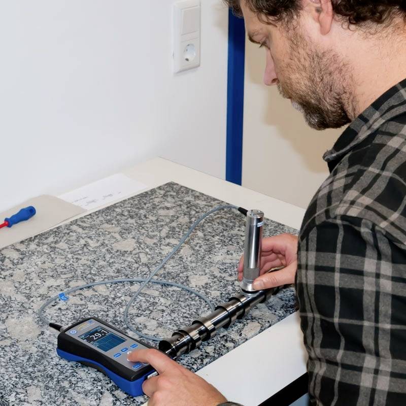 Muž měří napětí na kovovém spínacím prvku pomocí digitálního měřicího přístroje umístěného na stole.