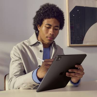 Osoba s kudrnatými vlasy sedí u stolu a píše perem na tablet. V pozadí je viditelná tabule.