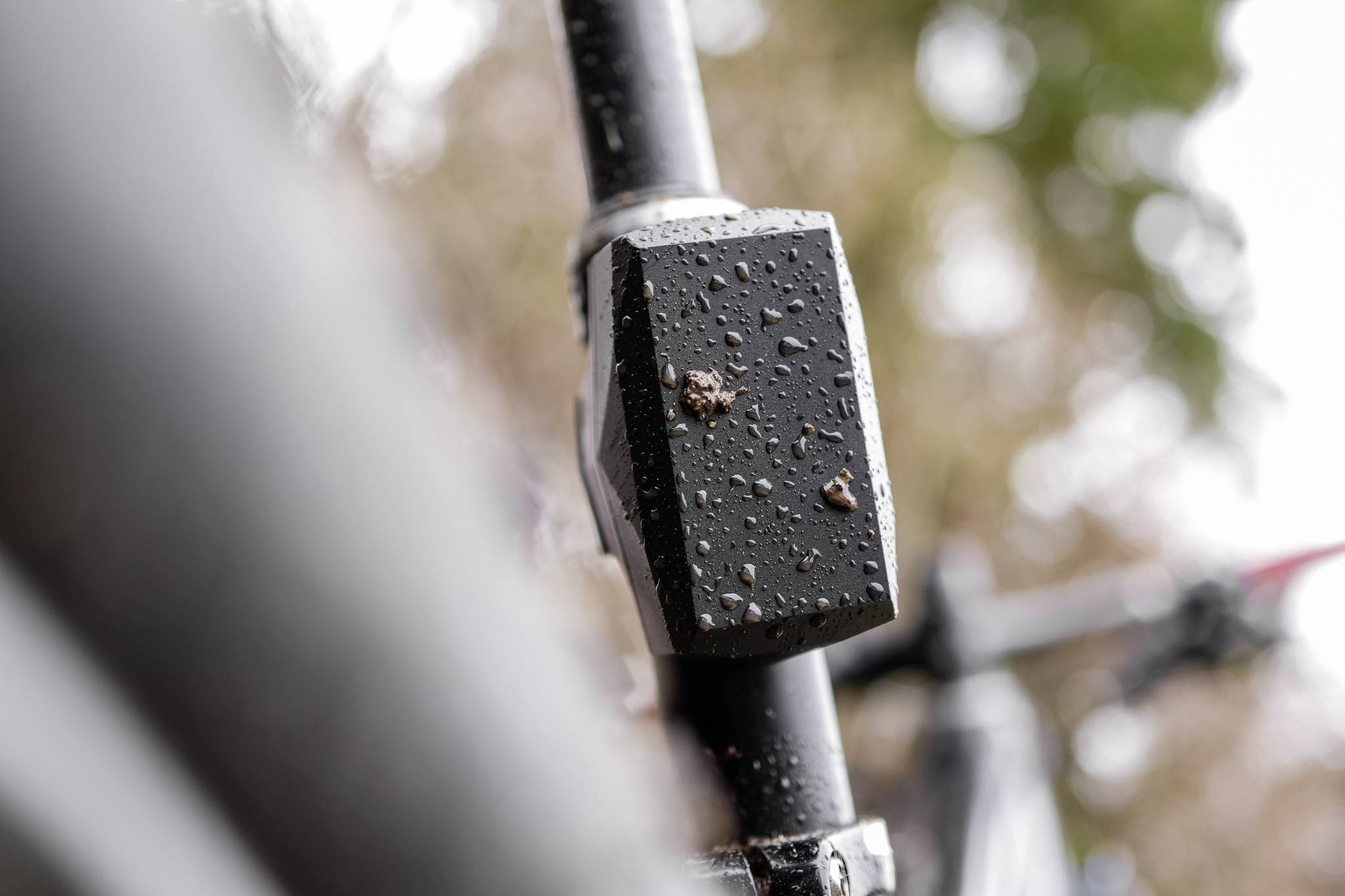 Detailní záběr cyklistické trubky s kapkami deště. V rozostřeném pozadí jsou viditelné stromy.