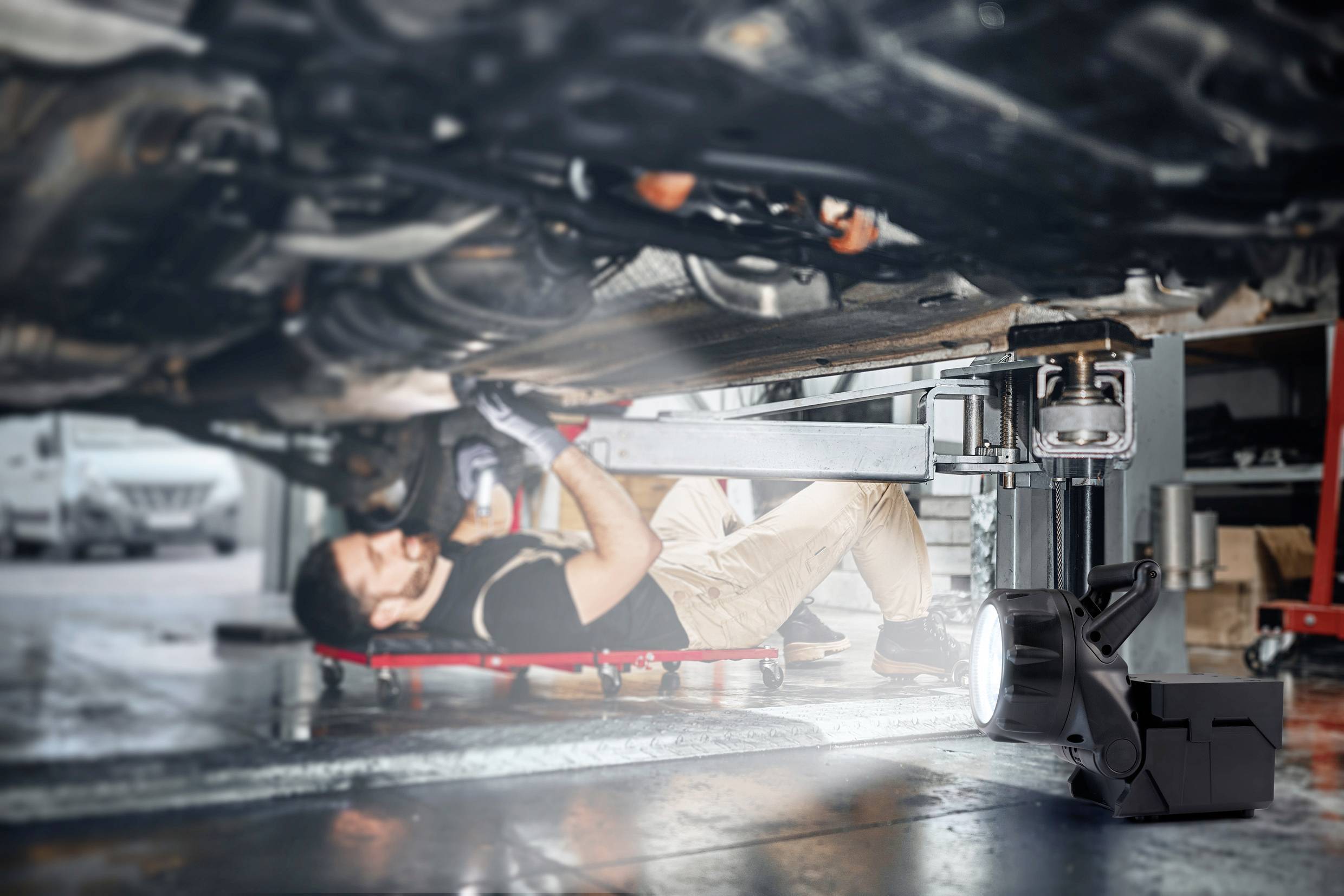 Mechanik leží pod autem v autoservisu, vedle něj je pracovní světlo, opravuje součásti vozidla.