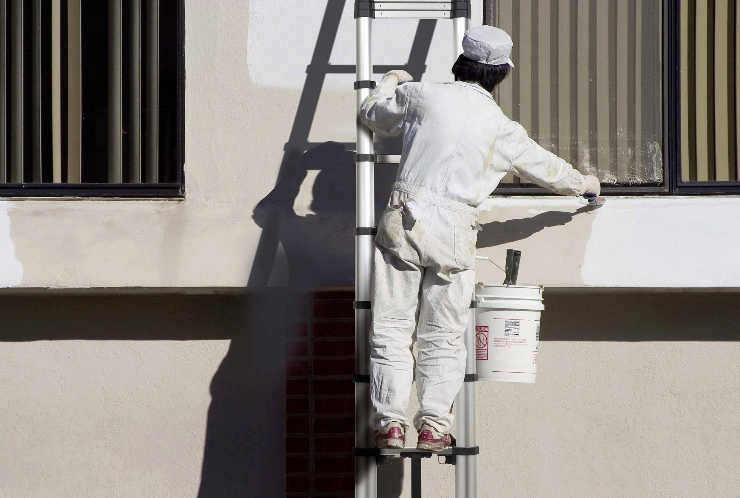 Malíř stojí na žebříku a natírá vnější stěnu budovy na bílo. Má na sobě ochranný oděv a vedle sebe kbelík s barvou.