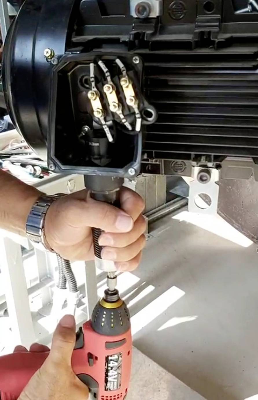 Osoba pracuje s červeným elektrickým vrtákem při údržbě elektromotoru. Je vidět několik kabelů a šroubů.