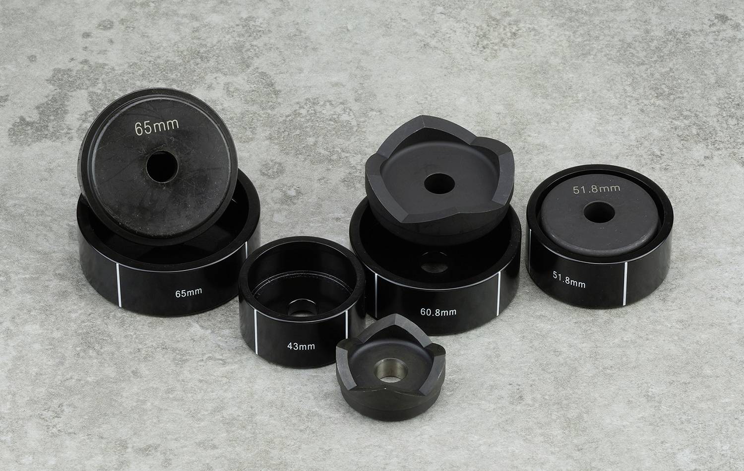 Sbírka černých kovových pouzder různých velikostí leží na šedém betonu. Jsou viditelné rozměry (51,8 mm, 43 mm, 65 mm).