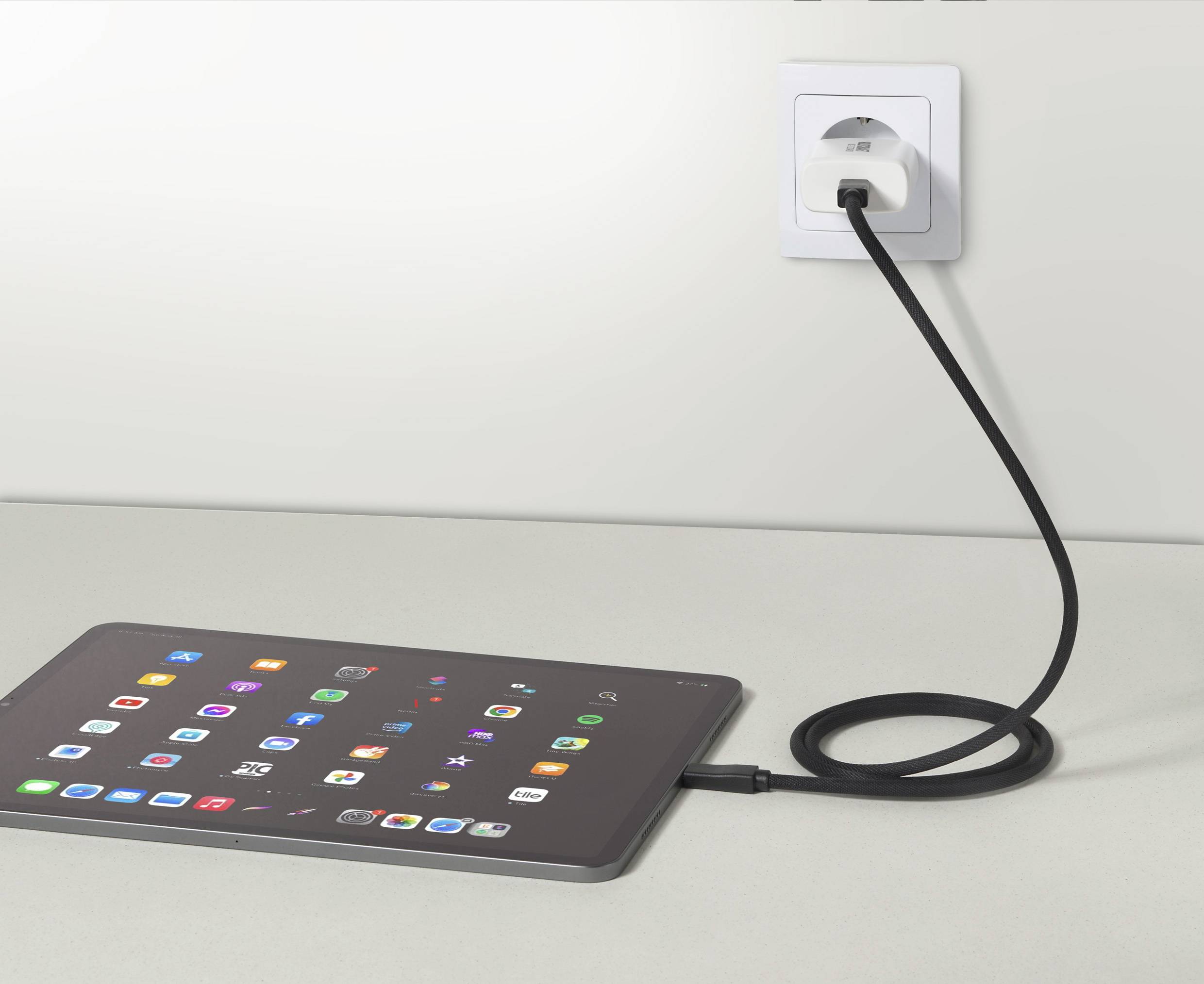 Tablet leží na stole a nabíjí se přes černý kabel zapojen do nástěnné zásuvky. Na obrazovce jsou viditelné různé ikony aplikací.
