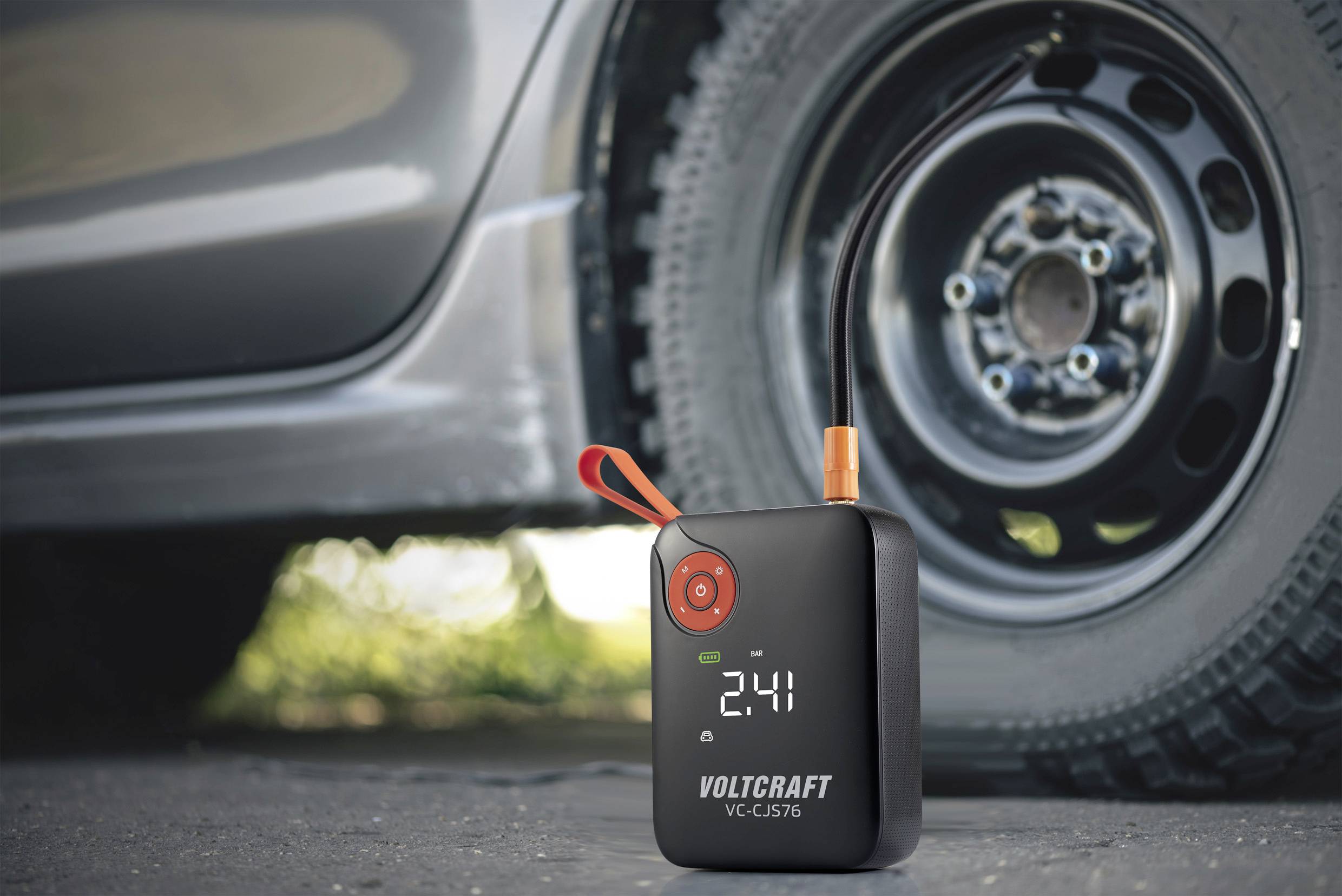 Přenosný kompresor na huštění pneumatik leží vedle autového kola a na displeji zobrazuje tlak '2.41', připravený k použití.