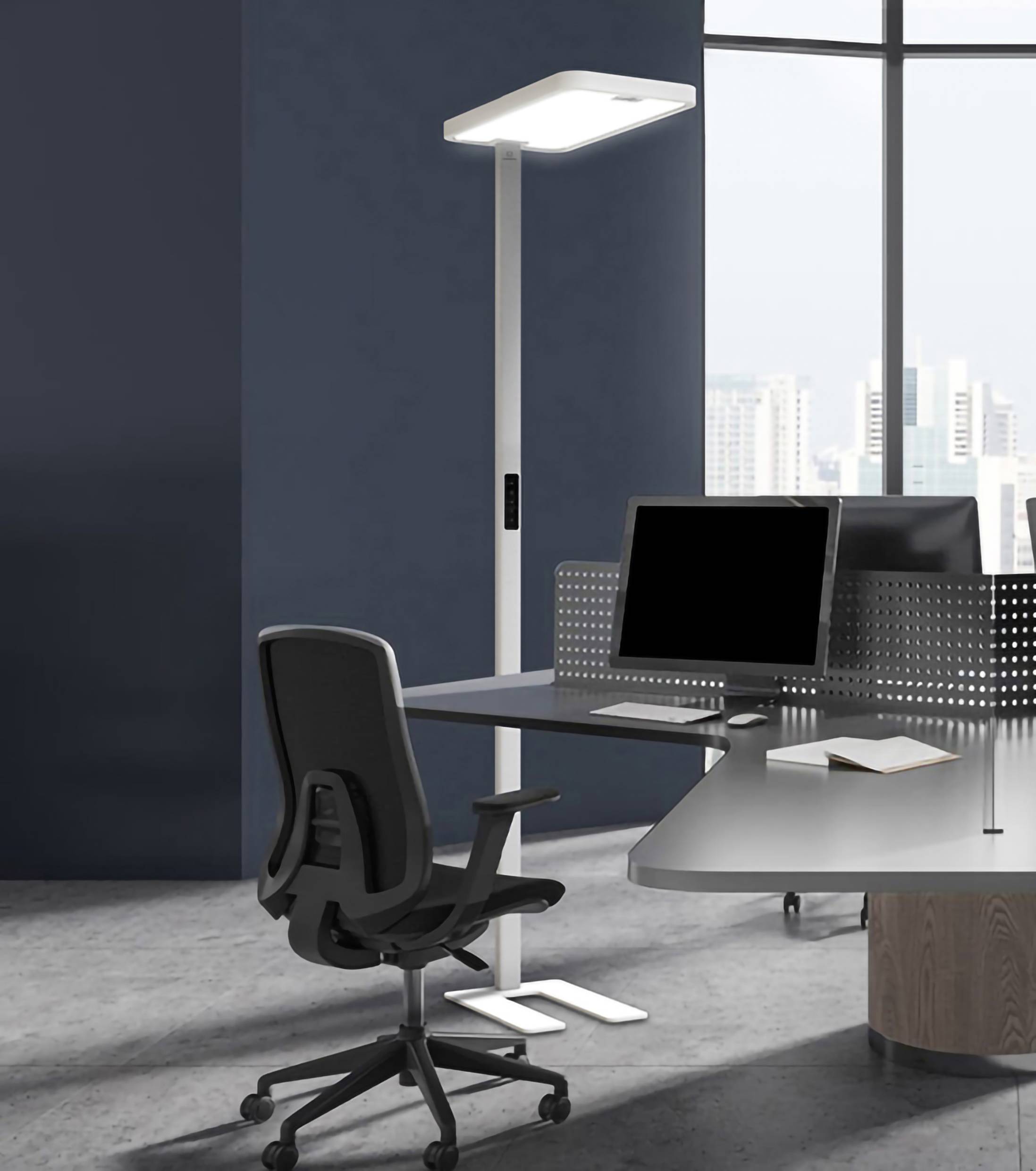 Moderní šedá kancelářská místnost s psacím stolem, černou kancelářskou židlí a velkým monitorem. V pozadí stojí vysoká stojací lampa před oknem s výhledem na město.