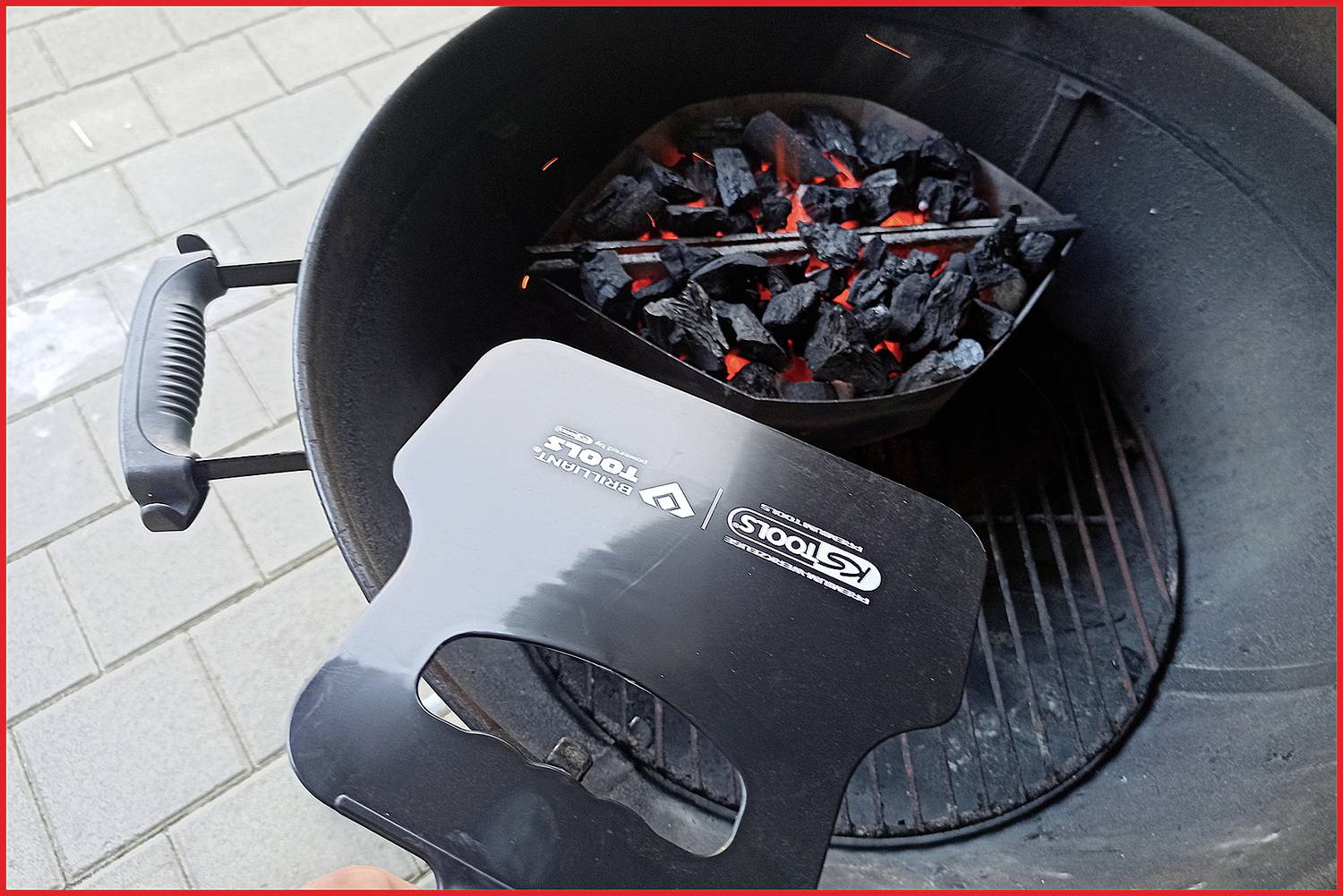 Gril s žhnoucím uhlím v uhelném koši. Víko grilu je přidržováno na okraji grilu, pravděpodobně za účelem jeho uzavření.