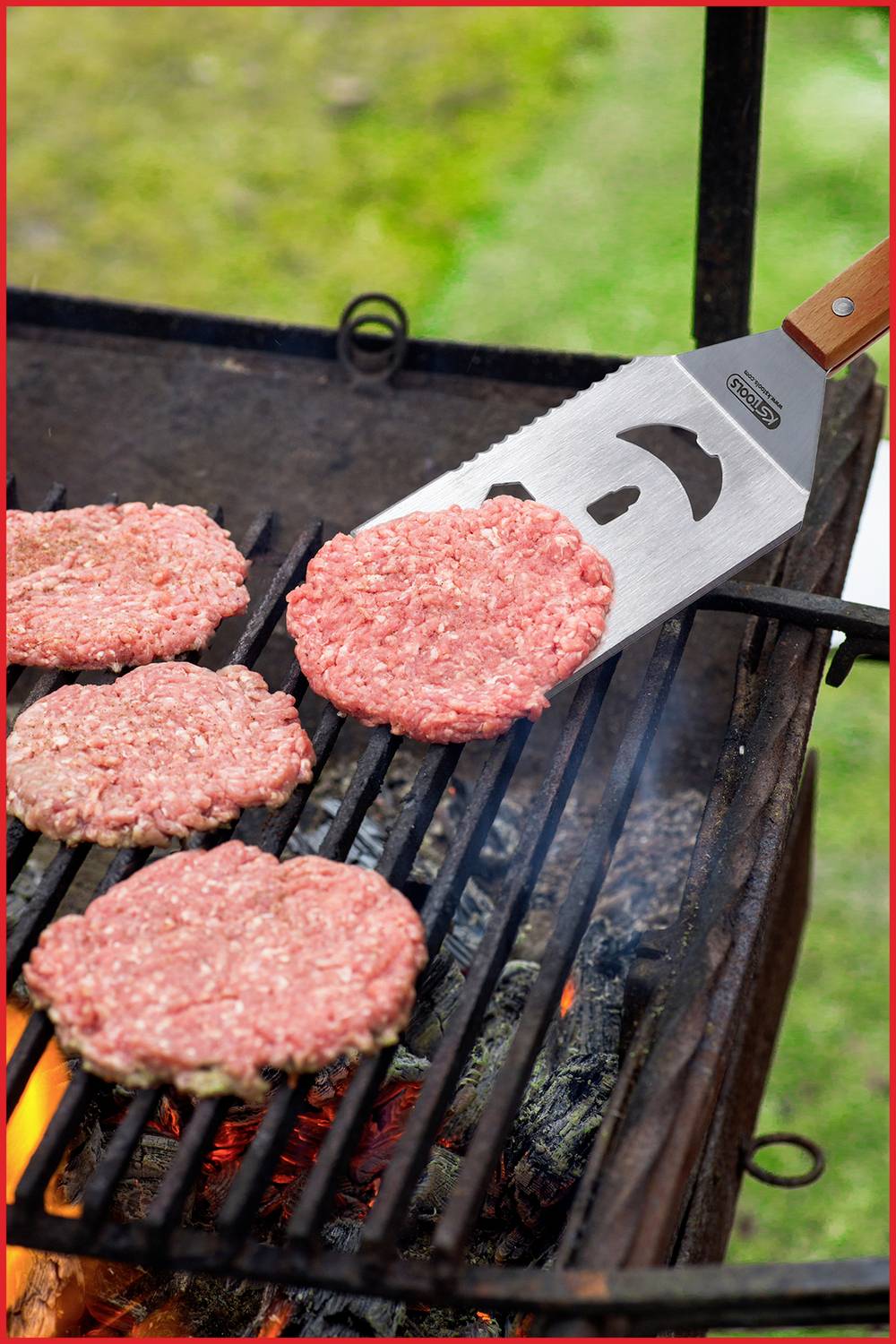 Čtyři syrové hamburgové placky jsou obraceny na grilu pomocí obracečky. Pod grilovacím roštem jsou viditelné plameny.