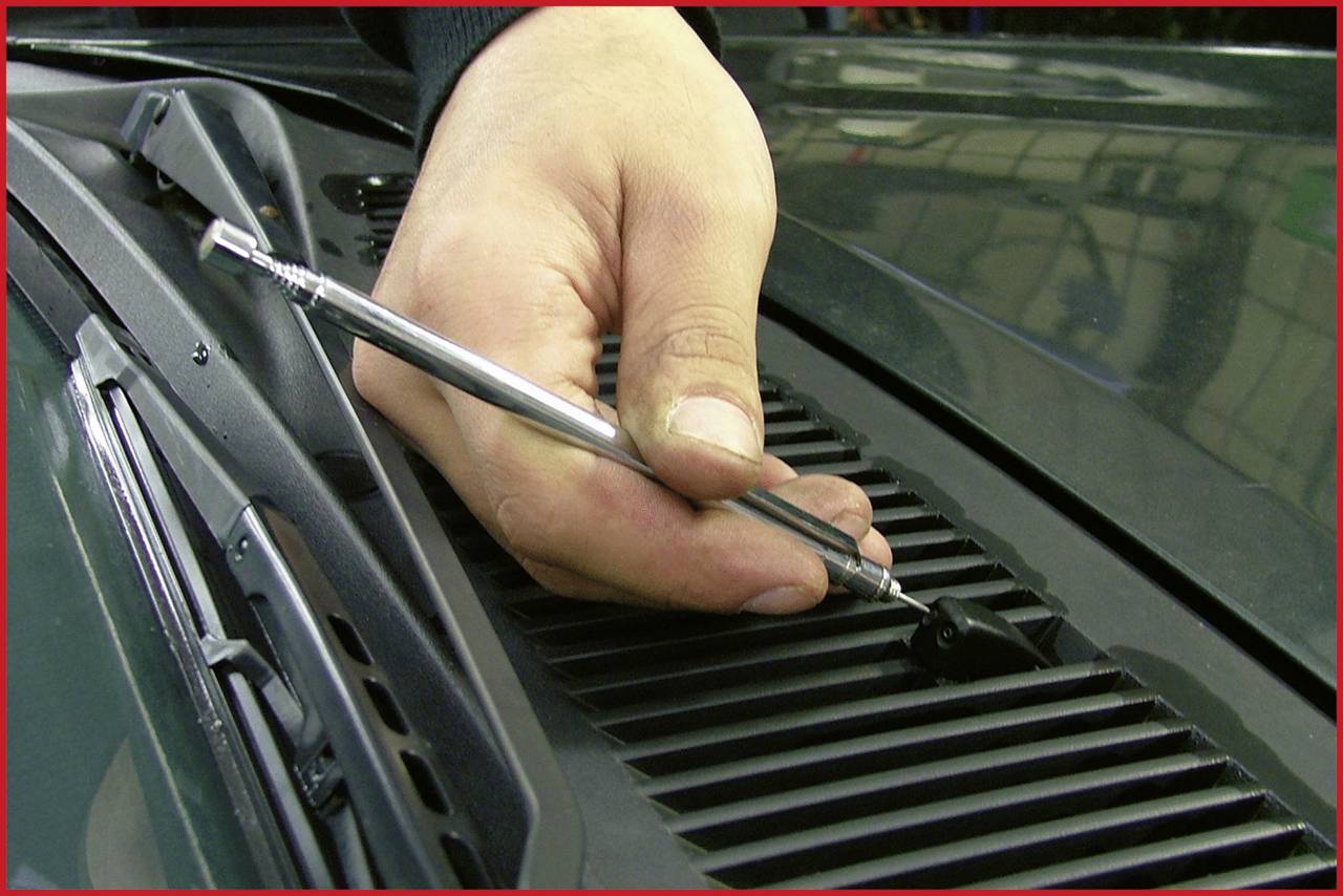 Jedna ruka drží stříbrný nástroj na mřížce ventilace automobilu. V pozadí je viditelný stín budovy.