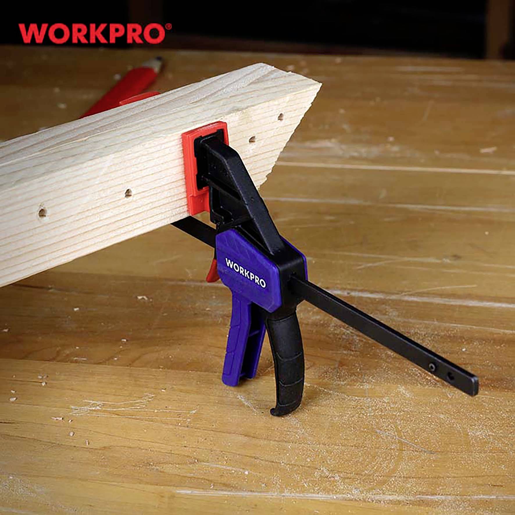 Modře-černé upínací nástroje drží dřevěnou desku na stole. Značka 'WORKPRO' je viditelná jak na nástroji, tak v celém obraze.