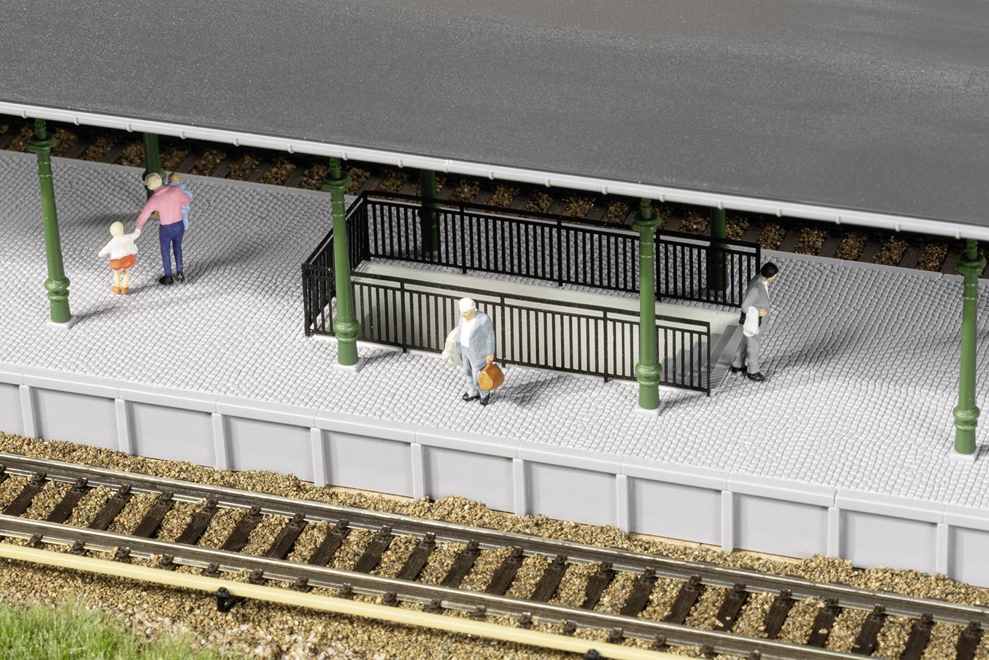 Miniaturní nástupiště se třemi figurkami: muž s dítětem, osoba sedící na lavičce a další člověk stojící a čekající.