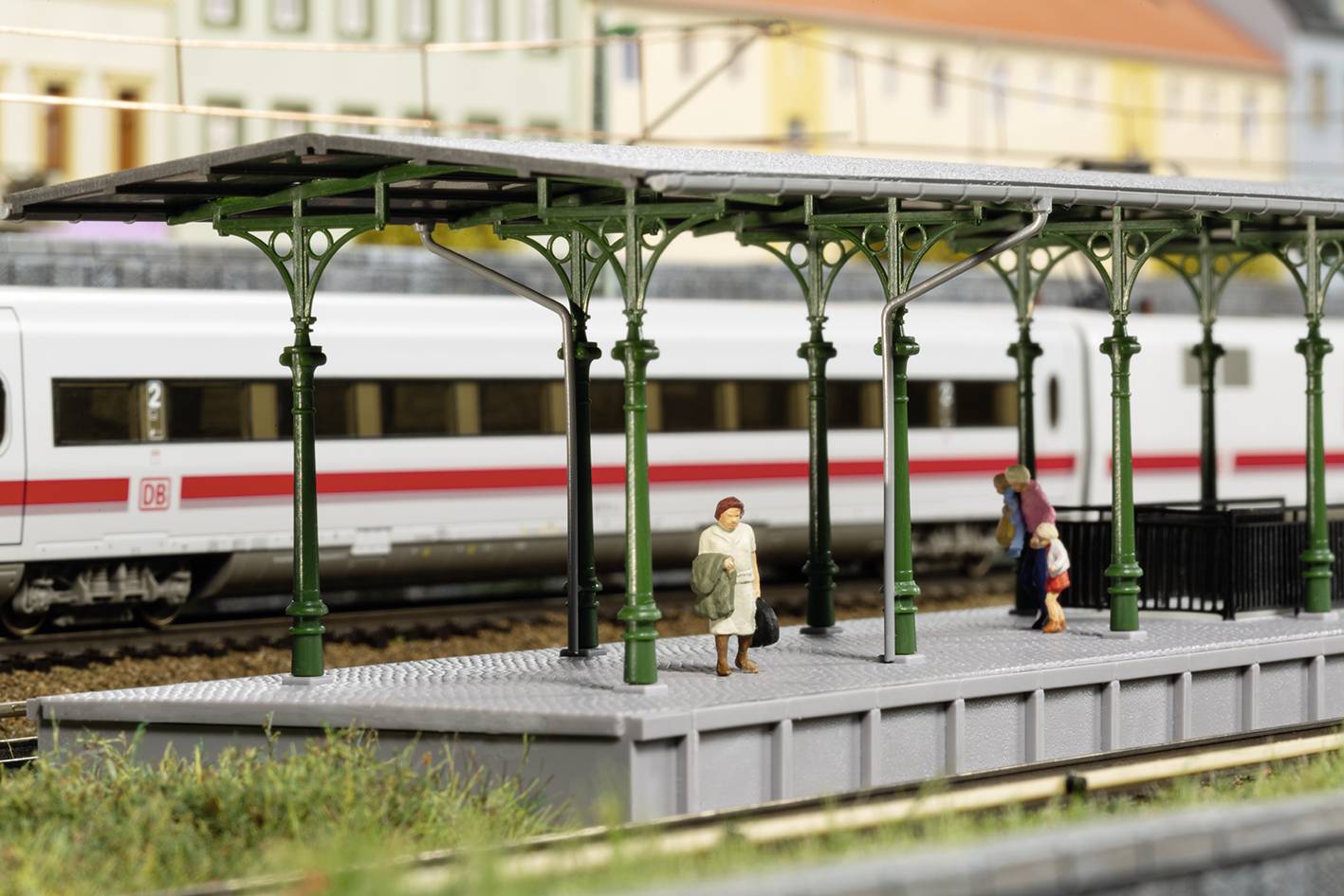 Miniaturní modelová železniční scéna s krytým nástupištěm, na kterém stojí dva lidé. V pozadí projíždí moderní vlak.
