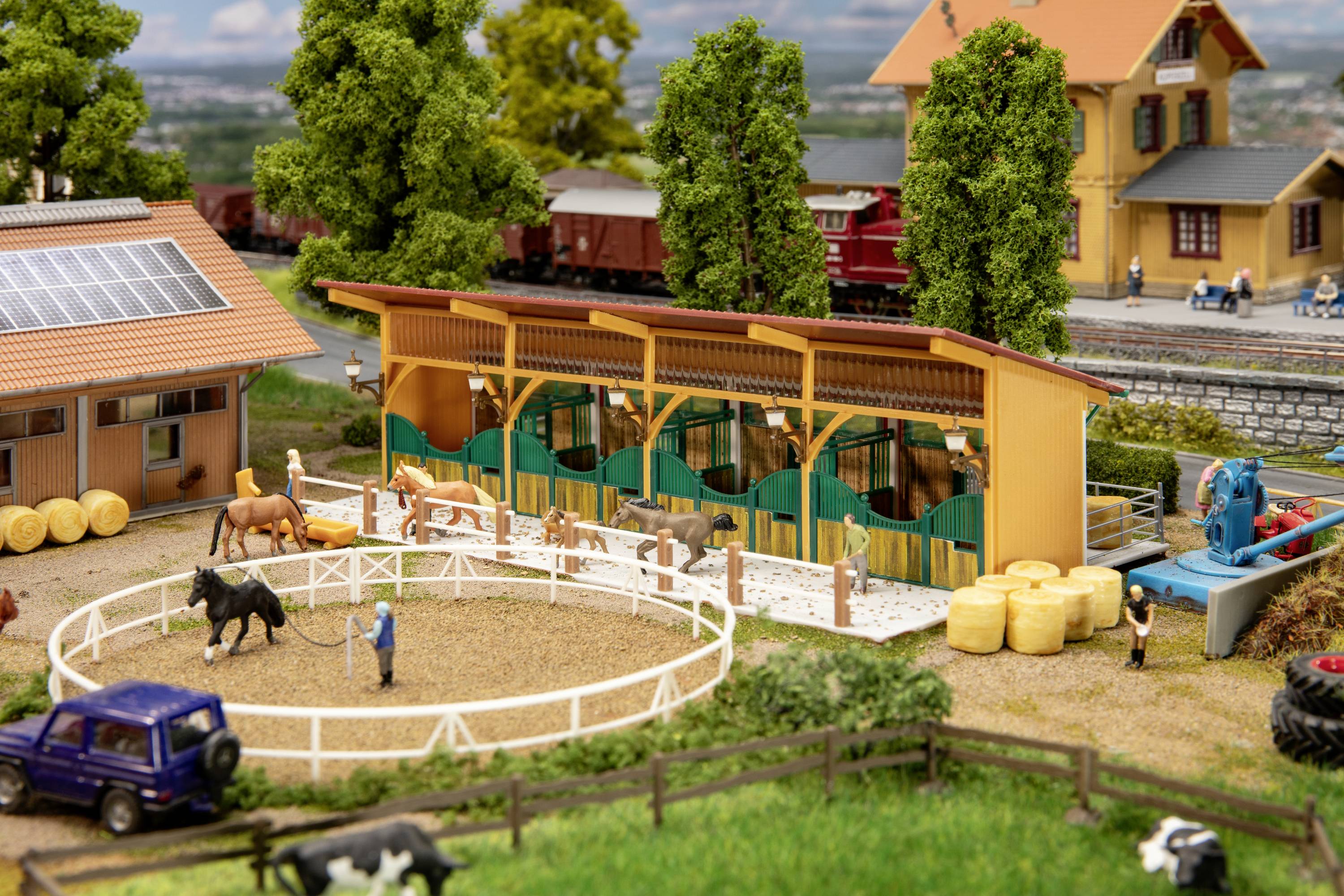 Miniaturnní krajina s koňskou stájí a malým tréninkovým prostorem. Trenér vede koně v kruhovém výběhu. Další koně jsou ve stáji.