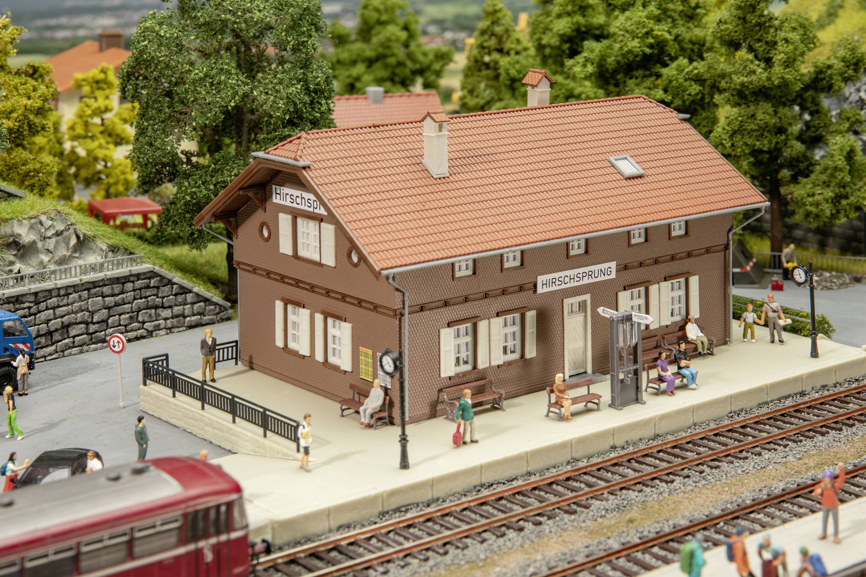 Modelové nádraží Hirschtal: Detailní miniatura nádraží s červenou cihlovou střechou, obklopená stromy a figurkami na nástupišti.
