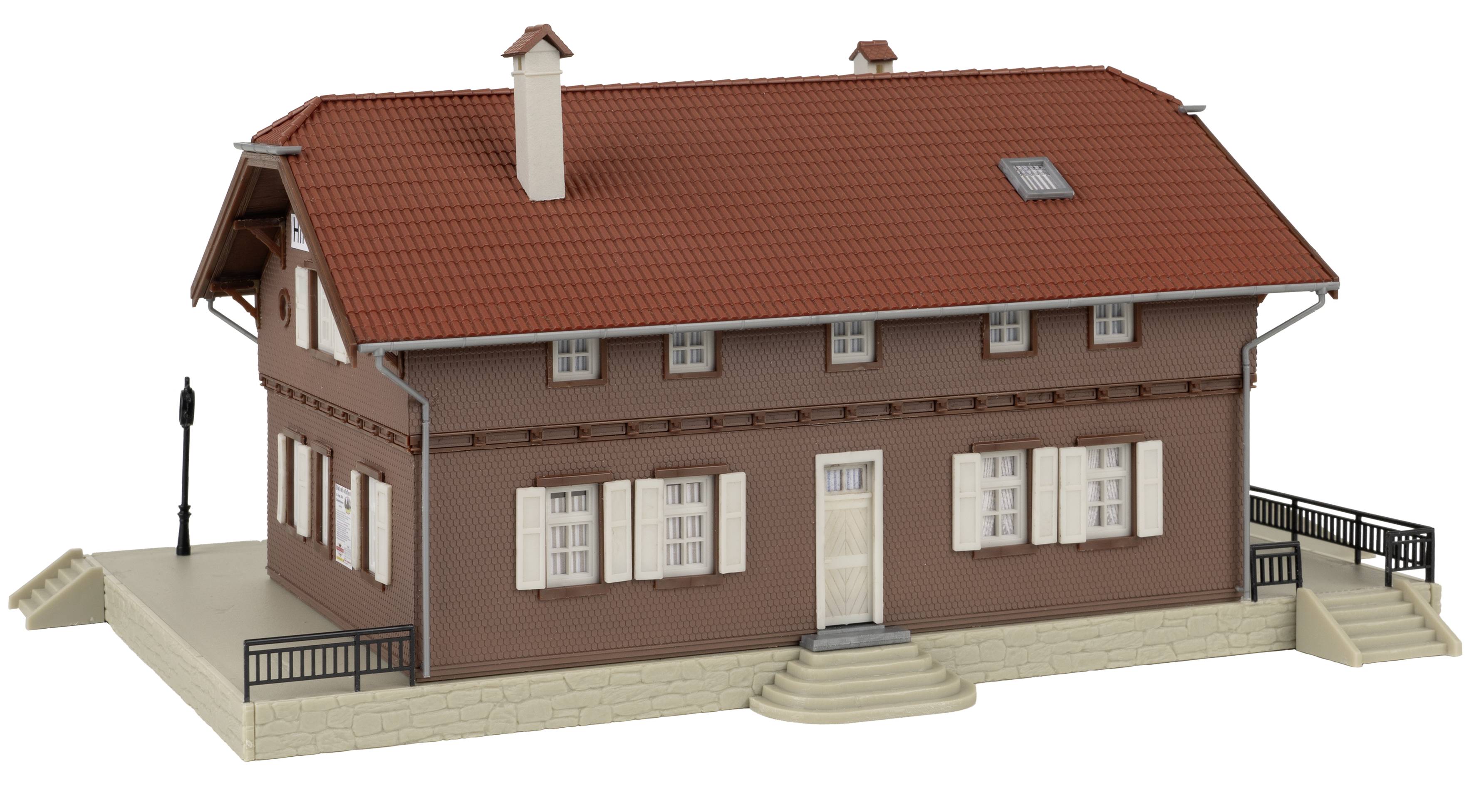 Malý model cihlového domu s červenou střechou a bílými okenicemi. Komíny a vstup se schodištěm jsou viditelné.