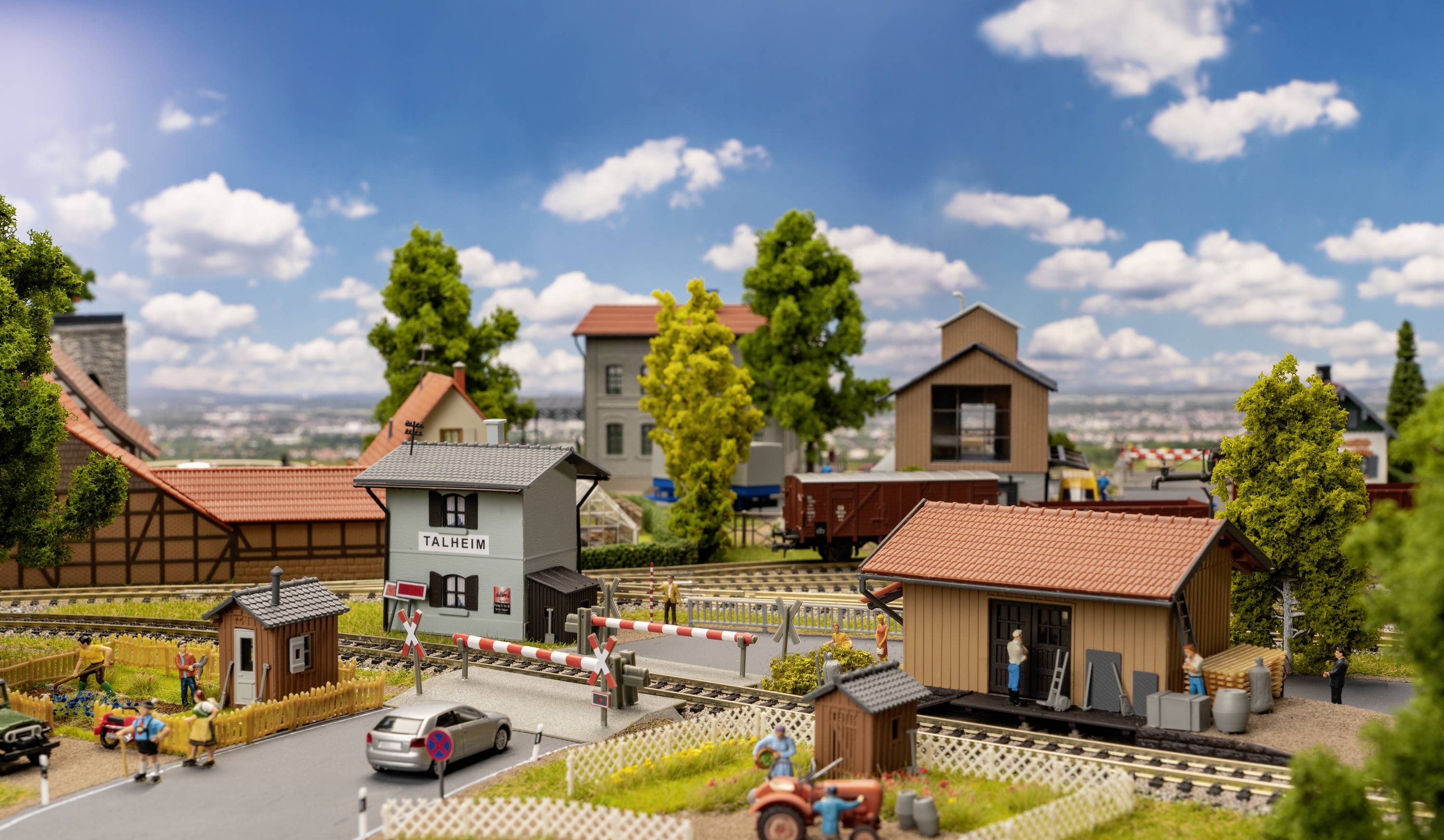Modelářská krajina s malými budovami, kolejemi, auty a stromy pod modrým nebem s mraky. Lidské figurky oživují scénu.