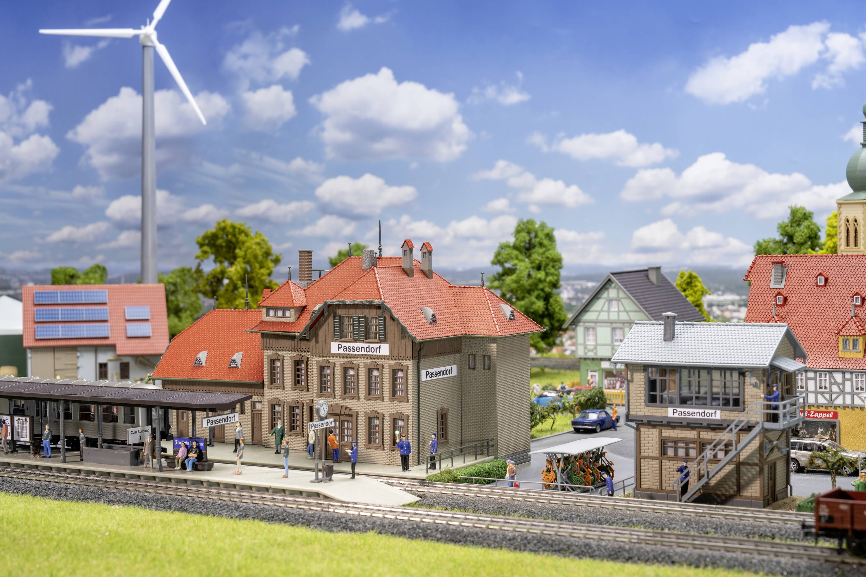Miniaturní model nádraží s větrnou elektrárnou v pozadí. Na nástupišti jsou vidět lidé. Kolem nádraží se nacházejí domy.