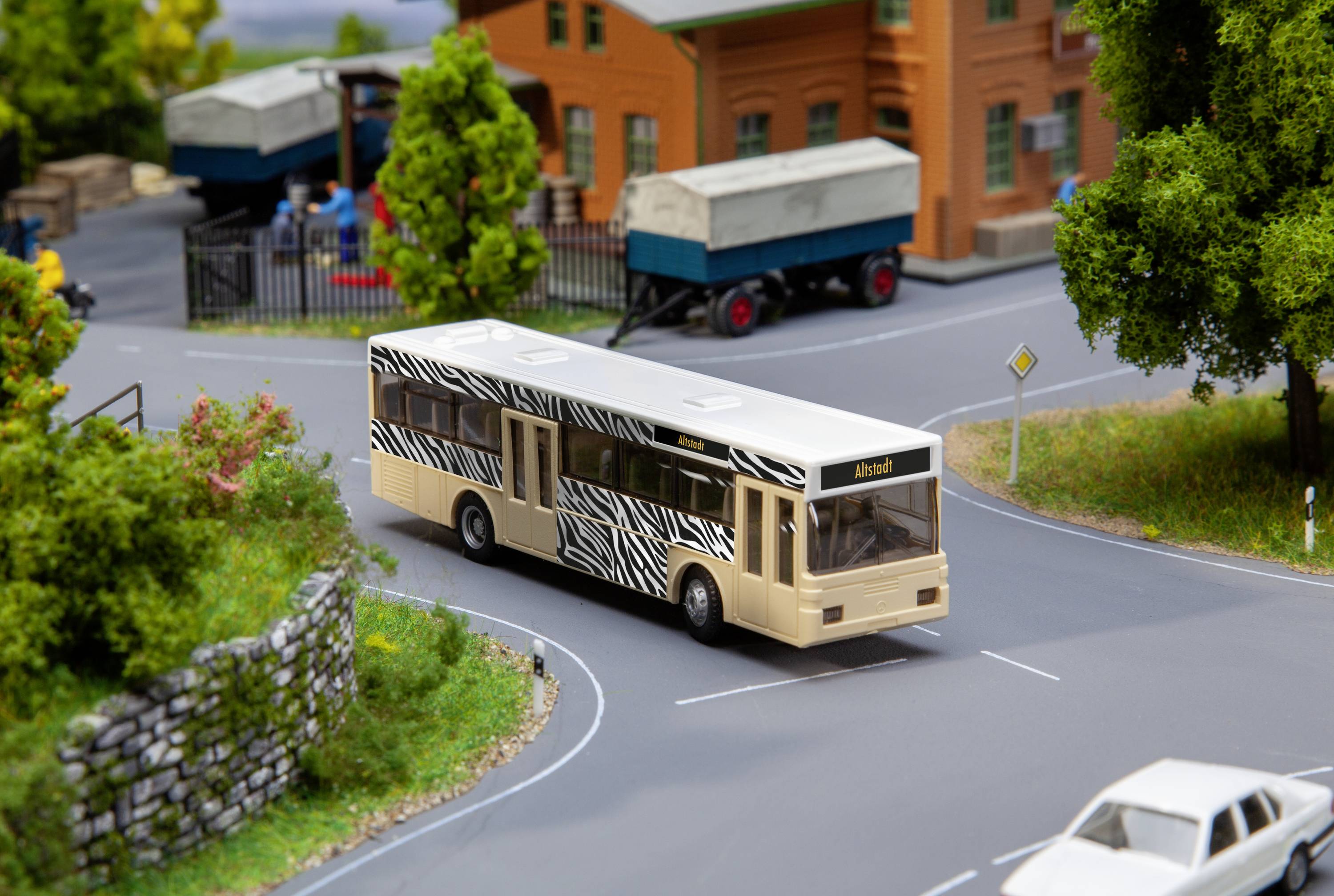 Modelový autobus s motivem zebry jede po miniaturní silnici. V pozadí jsou viditelné stromy, plot a nákladní automobil.