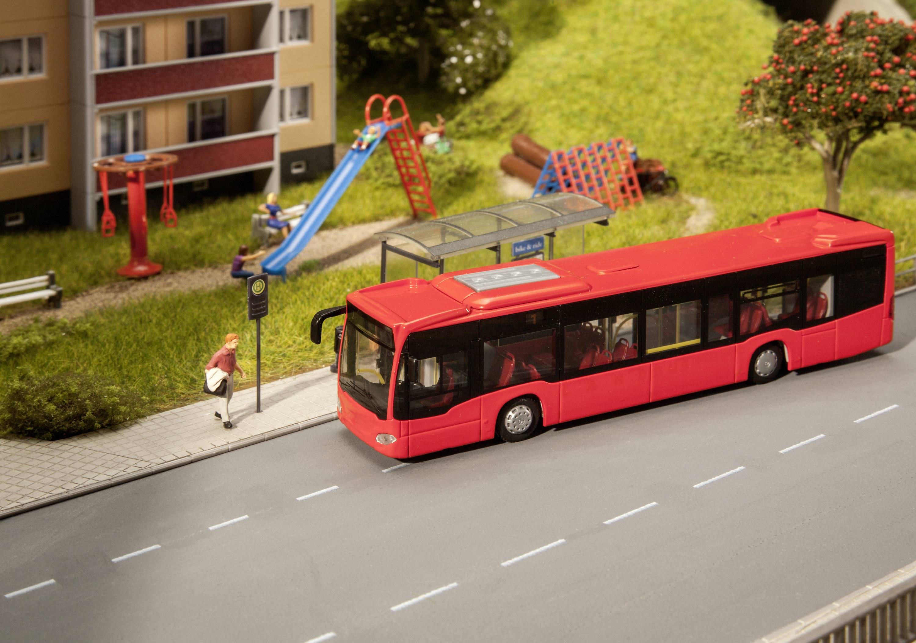 Červený autobus zastavuje na zastávce před bytovým domem. Jedna osoba se blíží k zastávce. V pozadí je vidět dětské hřiště.