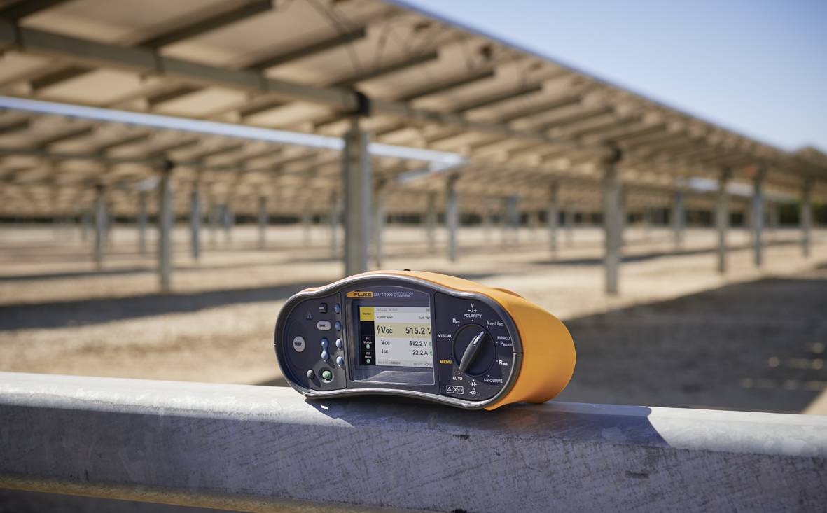 Digitální měřicí přístroj měří elektrické hodnoty pod rozsáhlou solární elektrárnou. Na pozadí jsou kovové struktury.