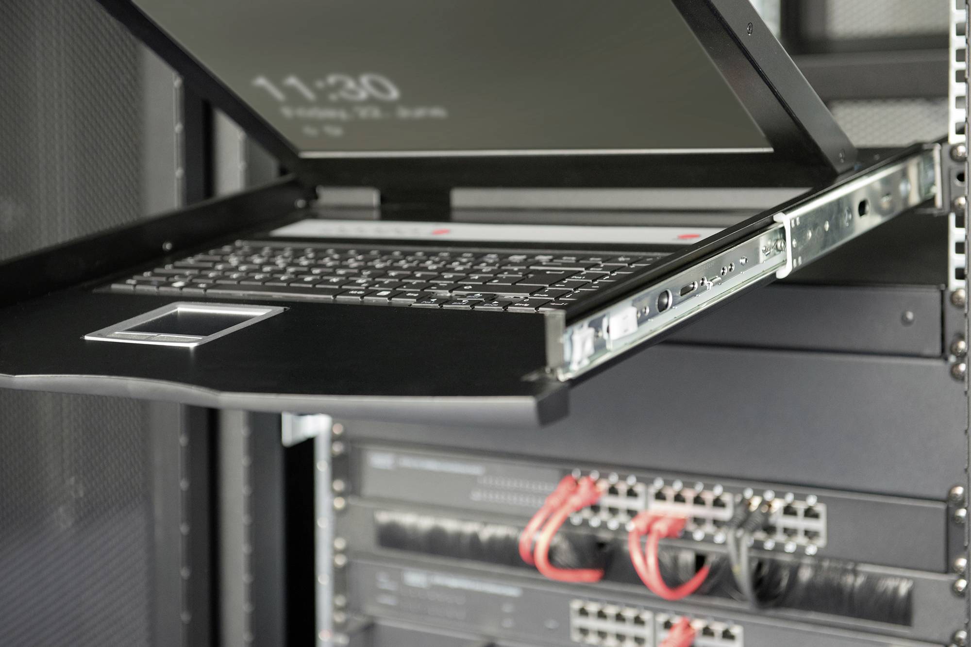 V serverovně je vidět otevřený laptop s displejem, který je namontovaný v rack liště. Pod ním jsou patrná síťová zařízení.