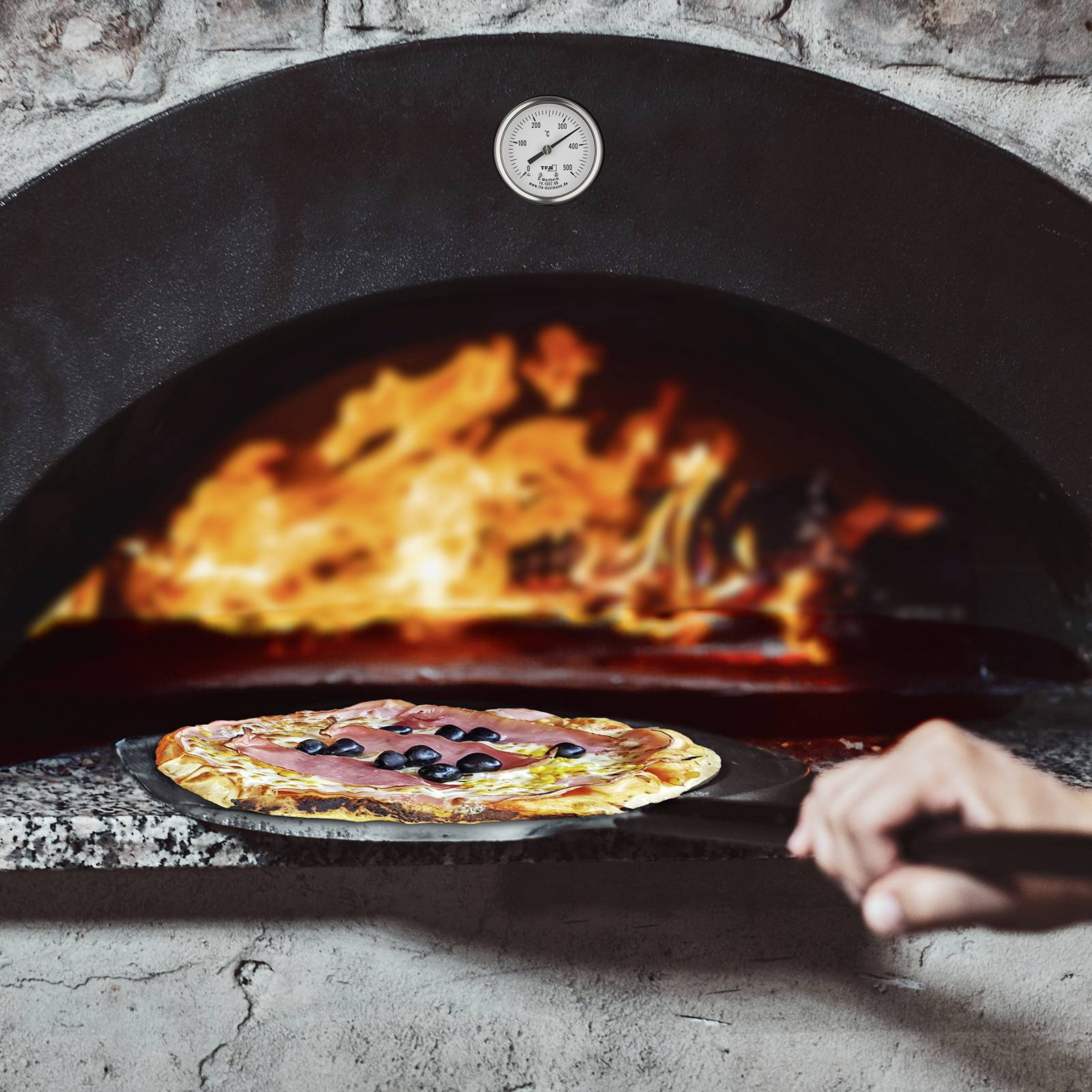 Jedna ruka drží lopatku s pizzou před tradičním kamenným dřevěným pecí, v níž hoří oheň.