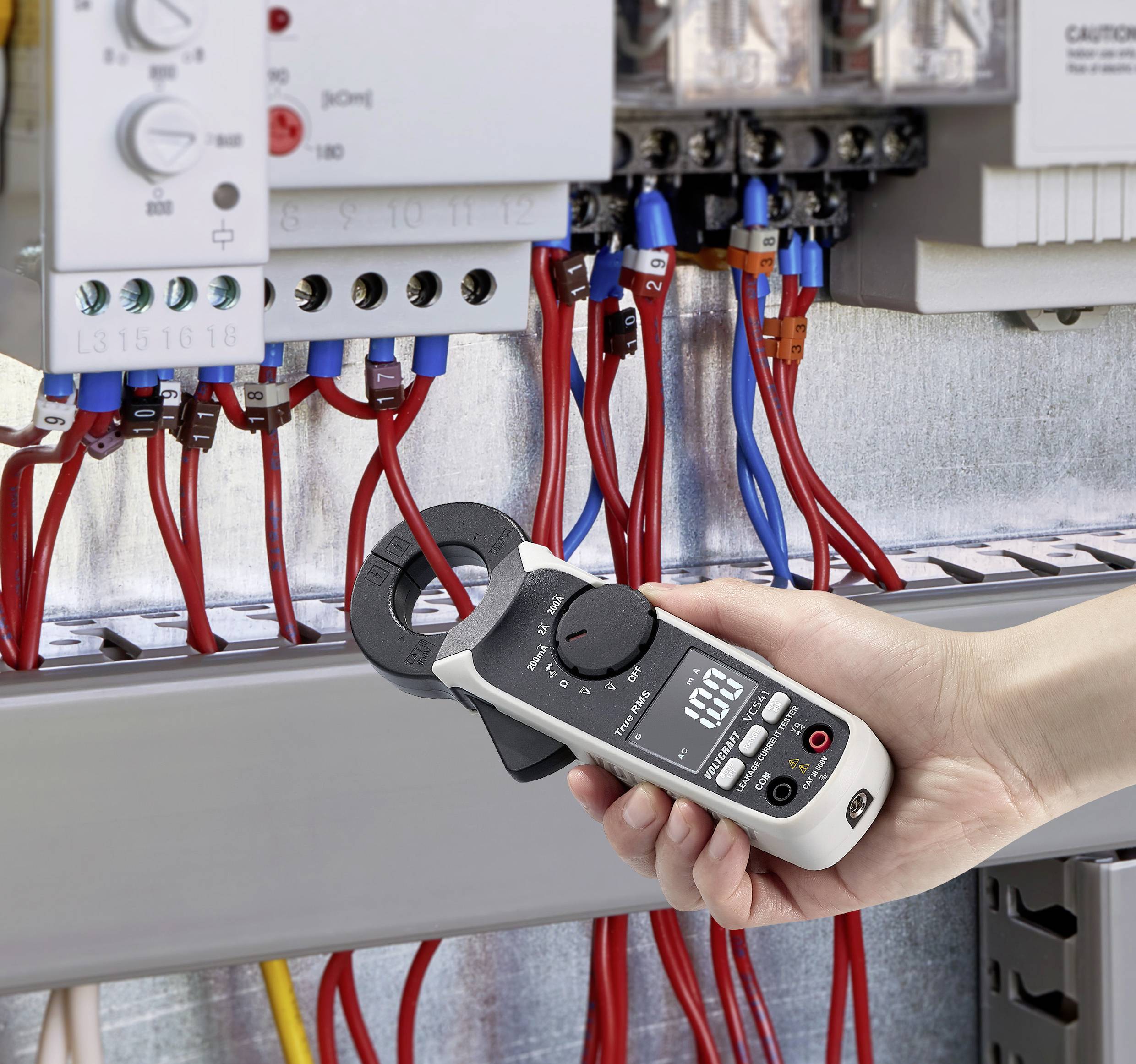 Osoba drží digitální multimetr před otevřenou elektrickou rozvodnou skříní a měří elektrické hodnoty pomocí červených a modrých měřicích kabelů.