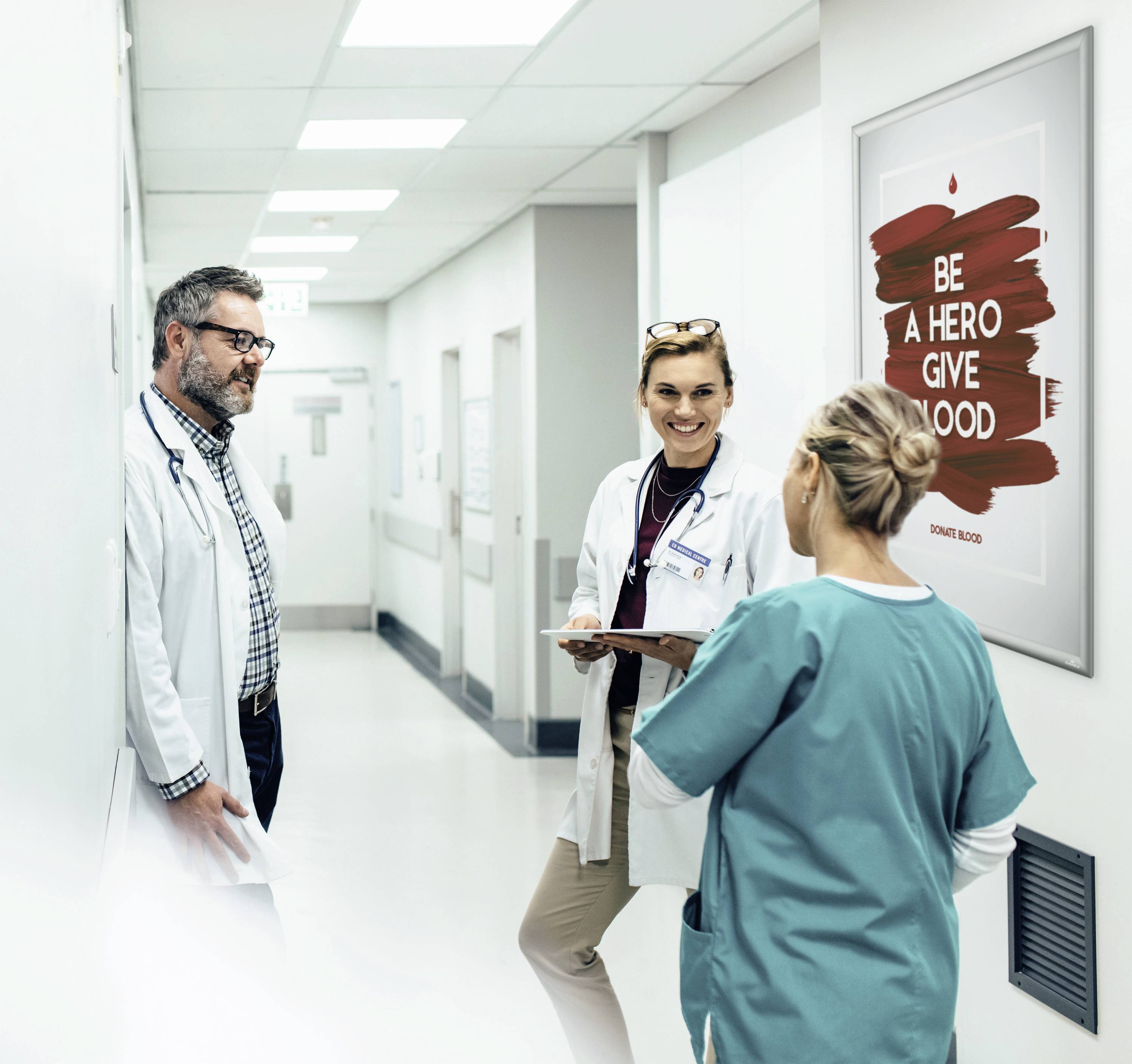 Tři zdravotničtí pracovníci se baví na nemocničním chodbě. Plakát na stěně hlásá: 'Buď hrdina, daruj krev'.