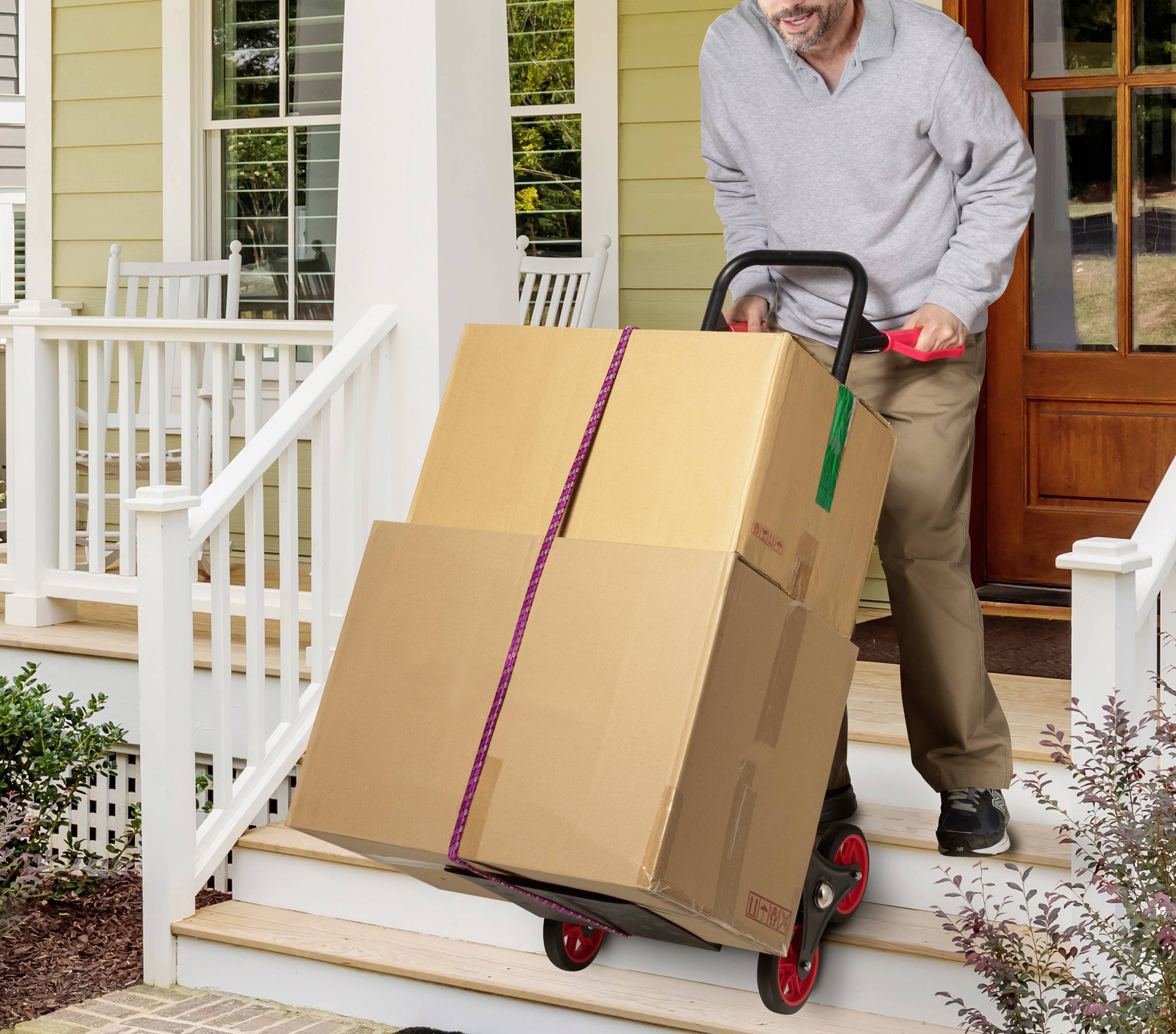 Muž veze na vozíku dva velké kartony do schodů verandy. V pozadí je viditelný dům.