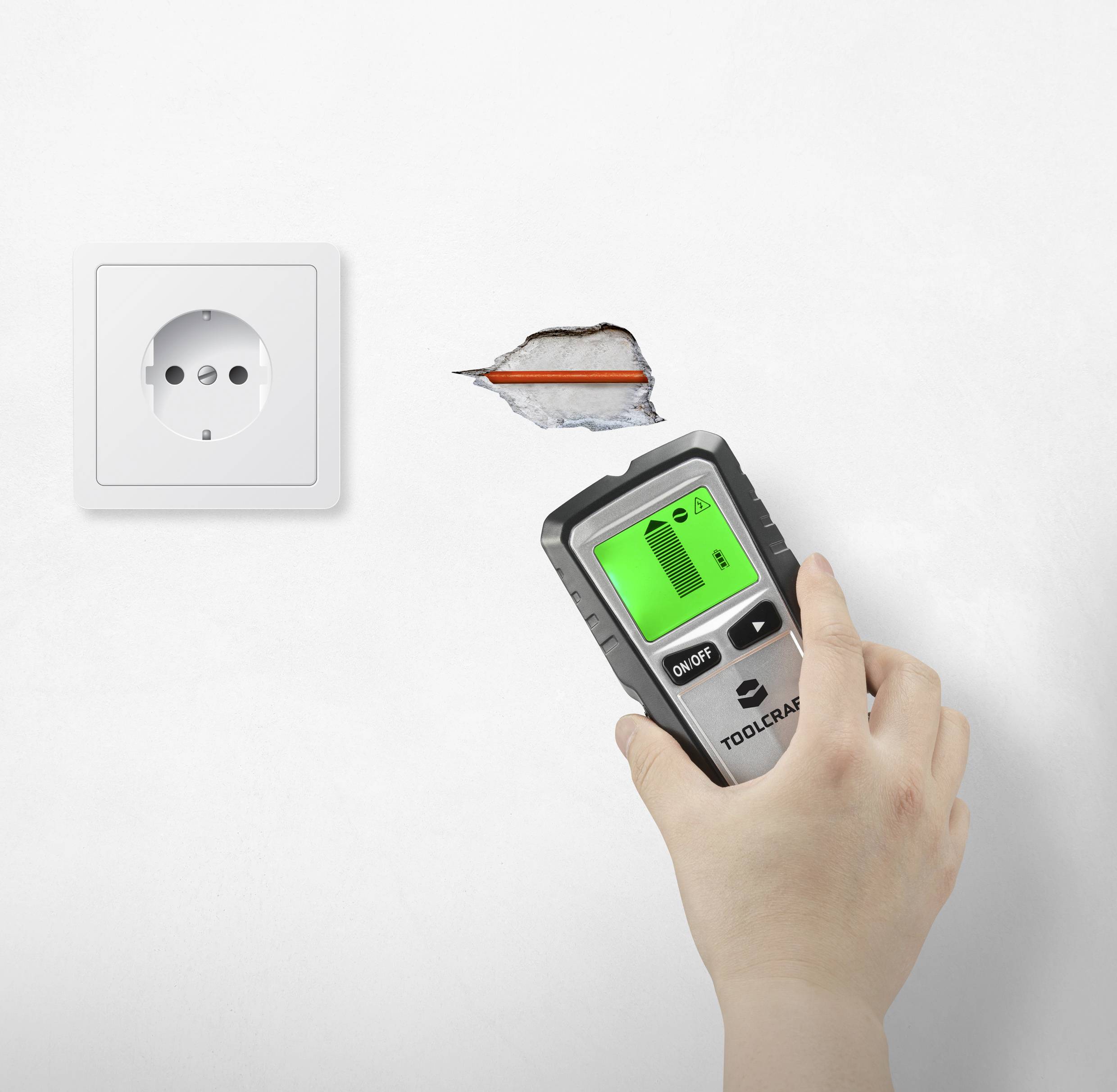 Jedna ruka drží elektronické detekční zařízení poblíž poškozené zdi s viditelnými vodiči vedle elektrické zásuvky.
