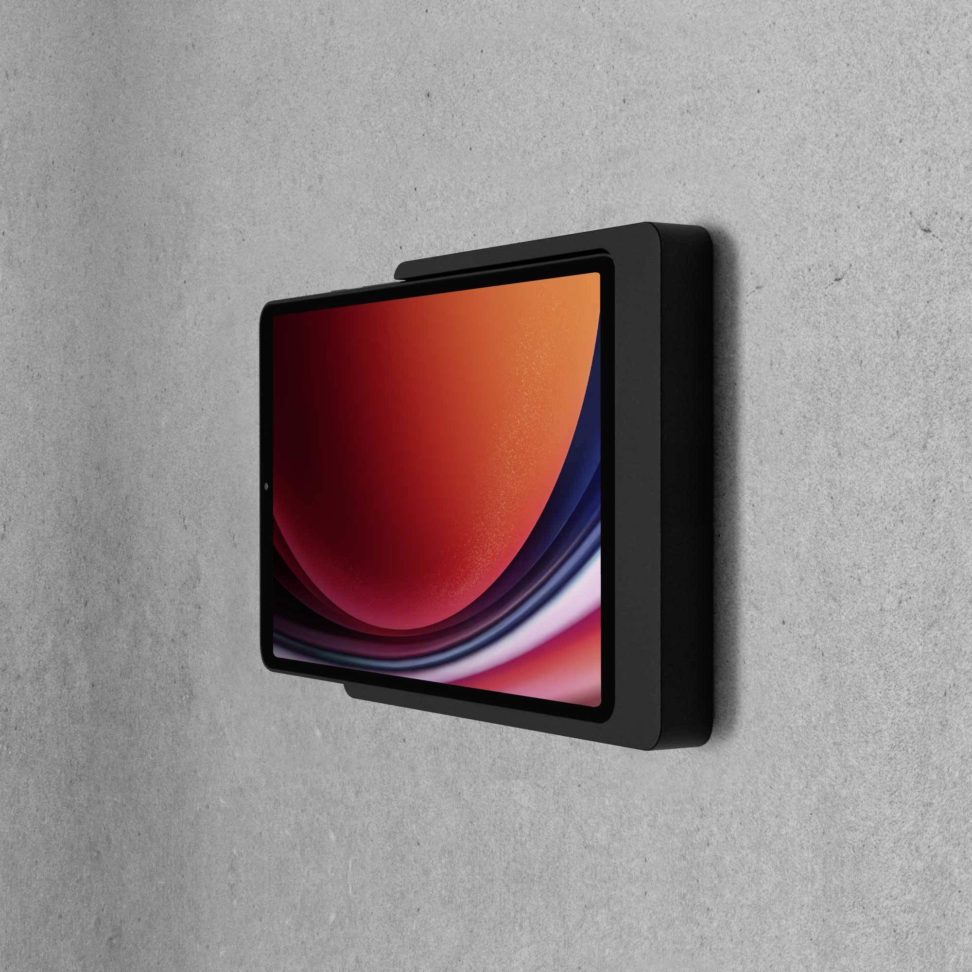 Tablet s červeným displejem visí vertikálně na šedé stěně.
