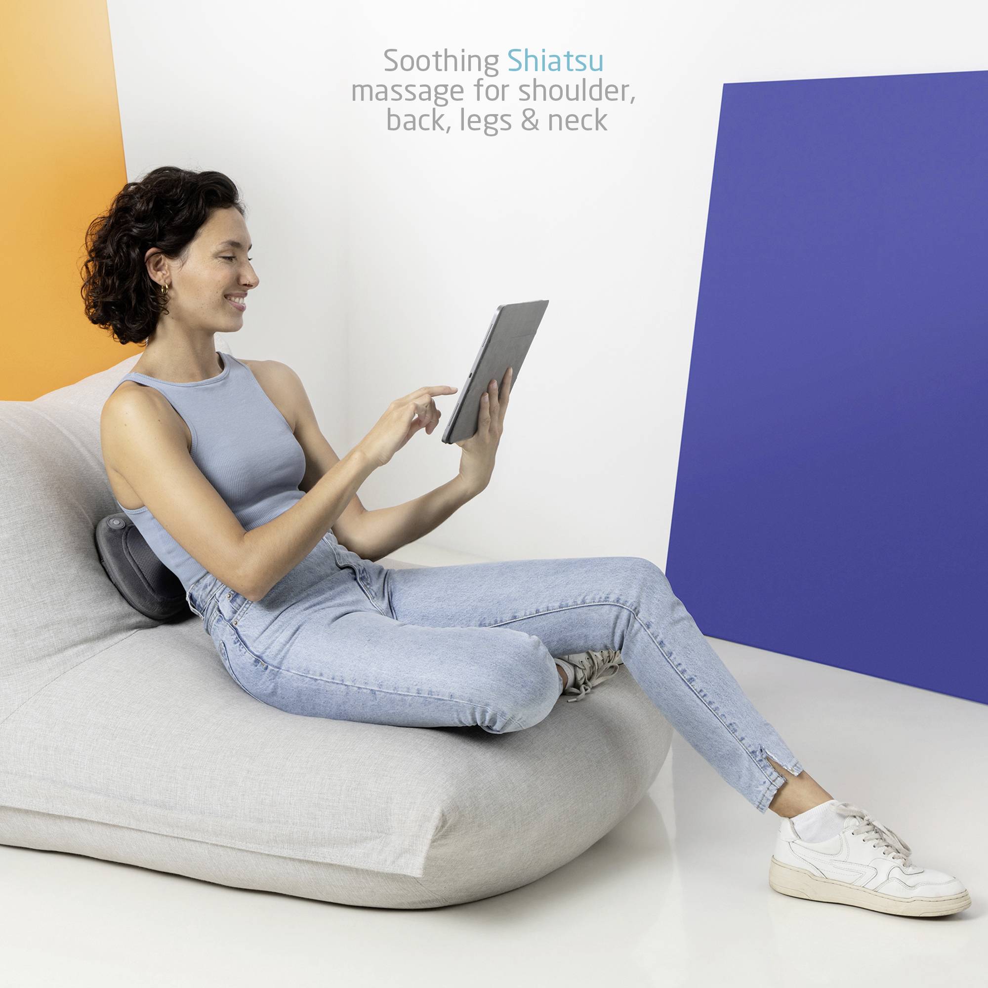 Žena sedí na pohovce, opírá se o masážní přístroj shiatsu a čte na tabletu. Místnost je světlá a moderně zařízená.