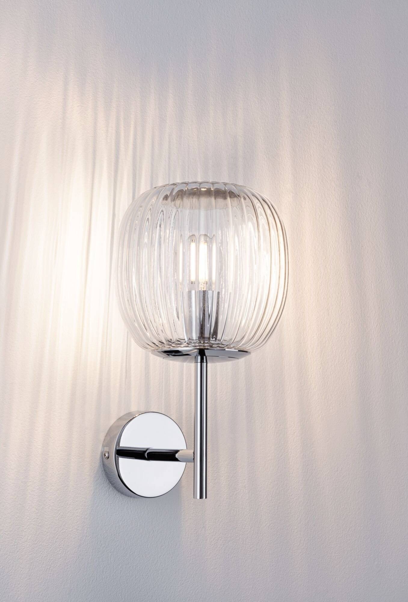 Nástěnná lampa s průhledným skleněným designem a kovovým držákem, která promítá teplé světlo na bílou stěnu.